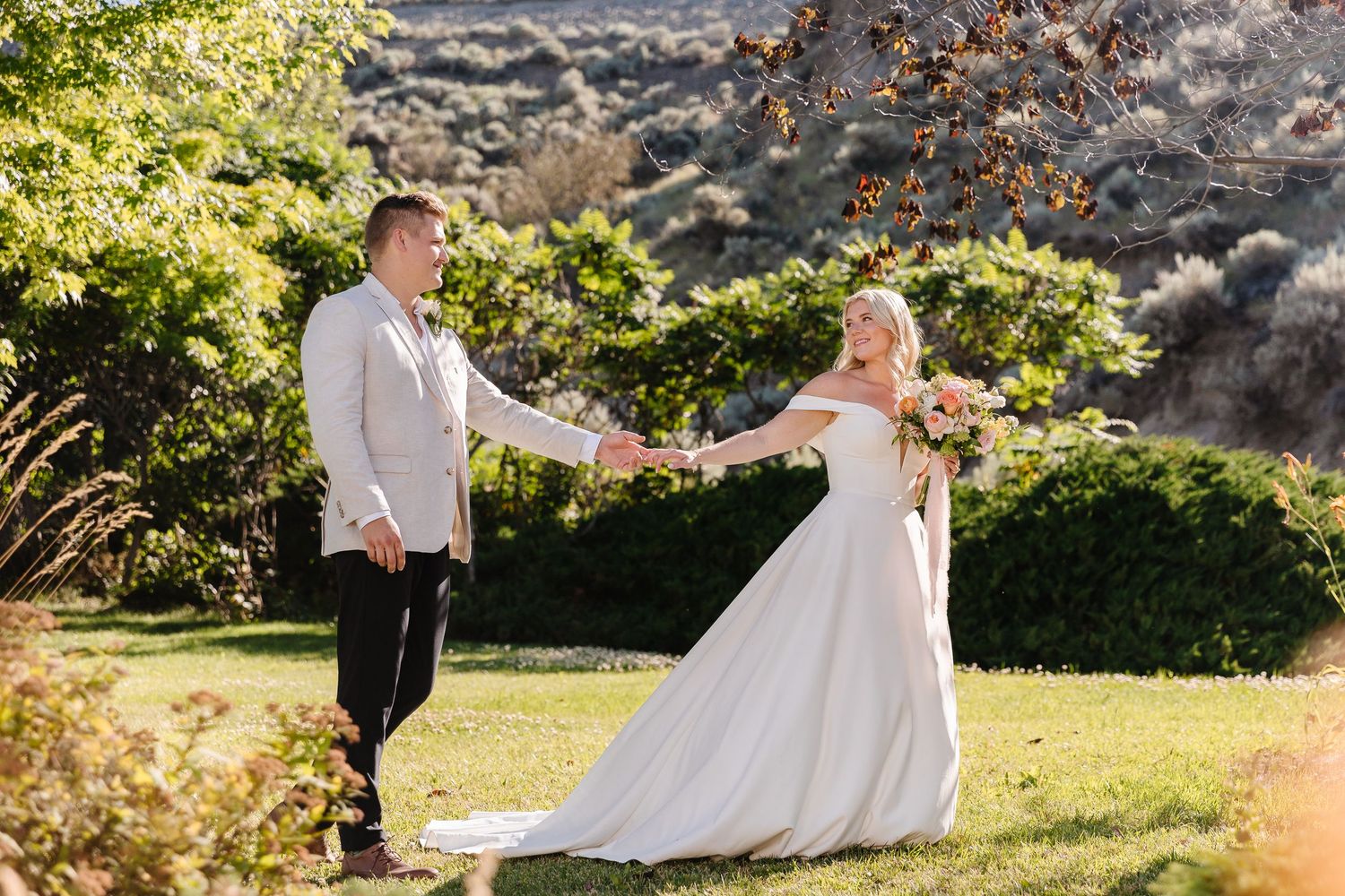 A couple in wedding attire dance and twirl together in a sunlit meadow surrounded by lush greenery.