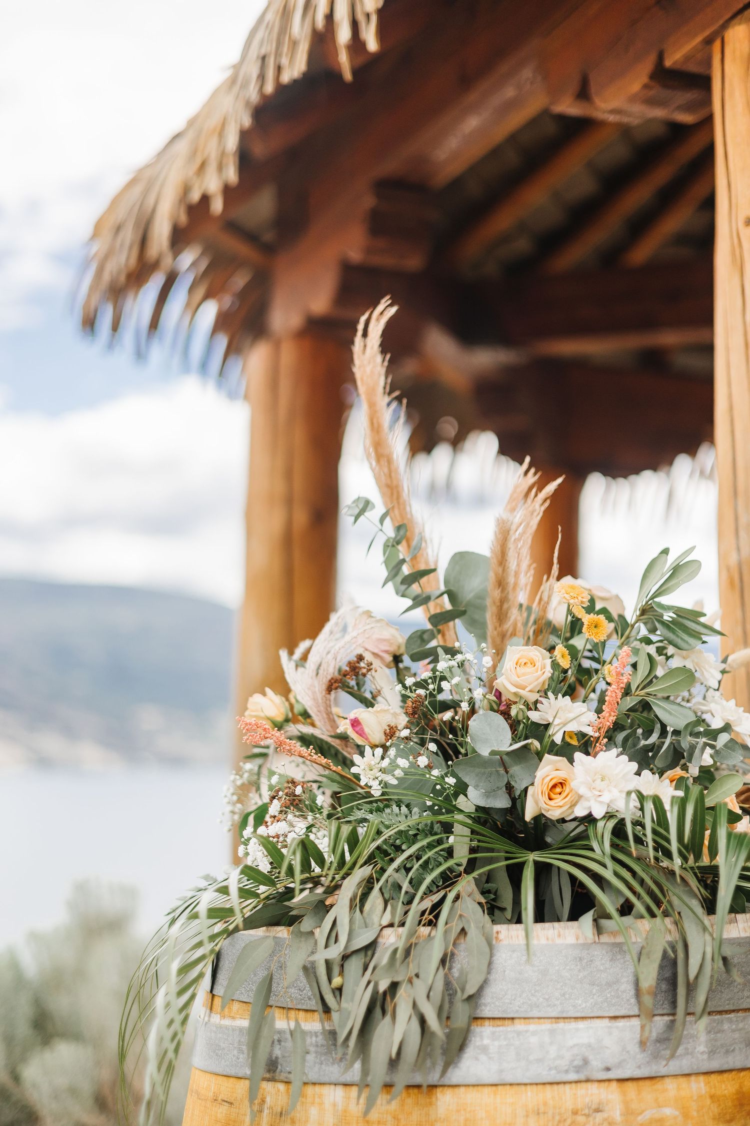 Rustic wedding floral arrangement with peach roses and eucalyptus displayed on a wooden barrel under a thatched roof.