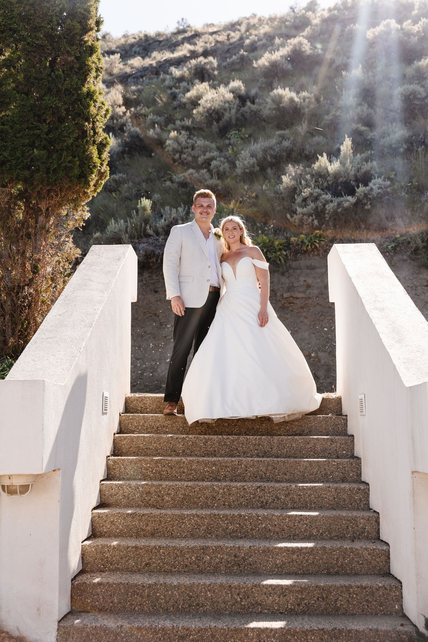 A couple in wedding attire descends a white stucco staircase with sunlight streaming through the trees.