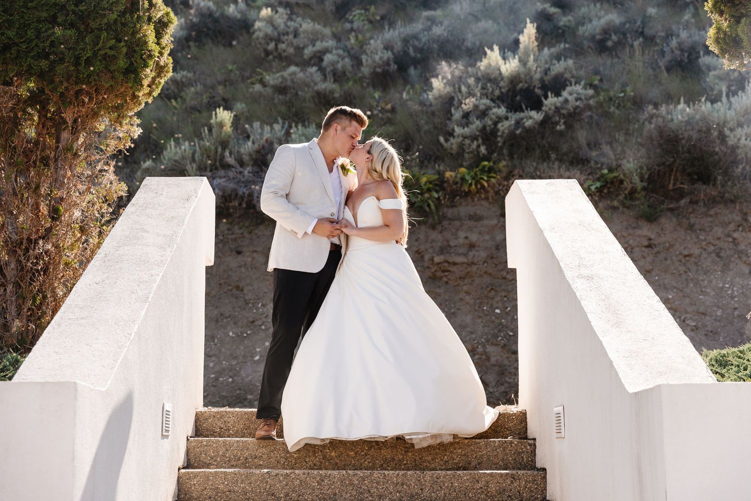 A couple in wedding attire embraces on stone steps surrounded by lush greenery and natural lighting.