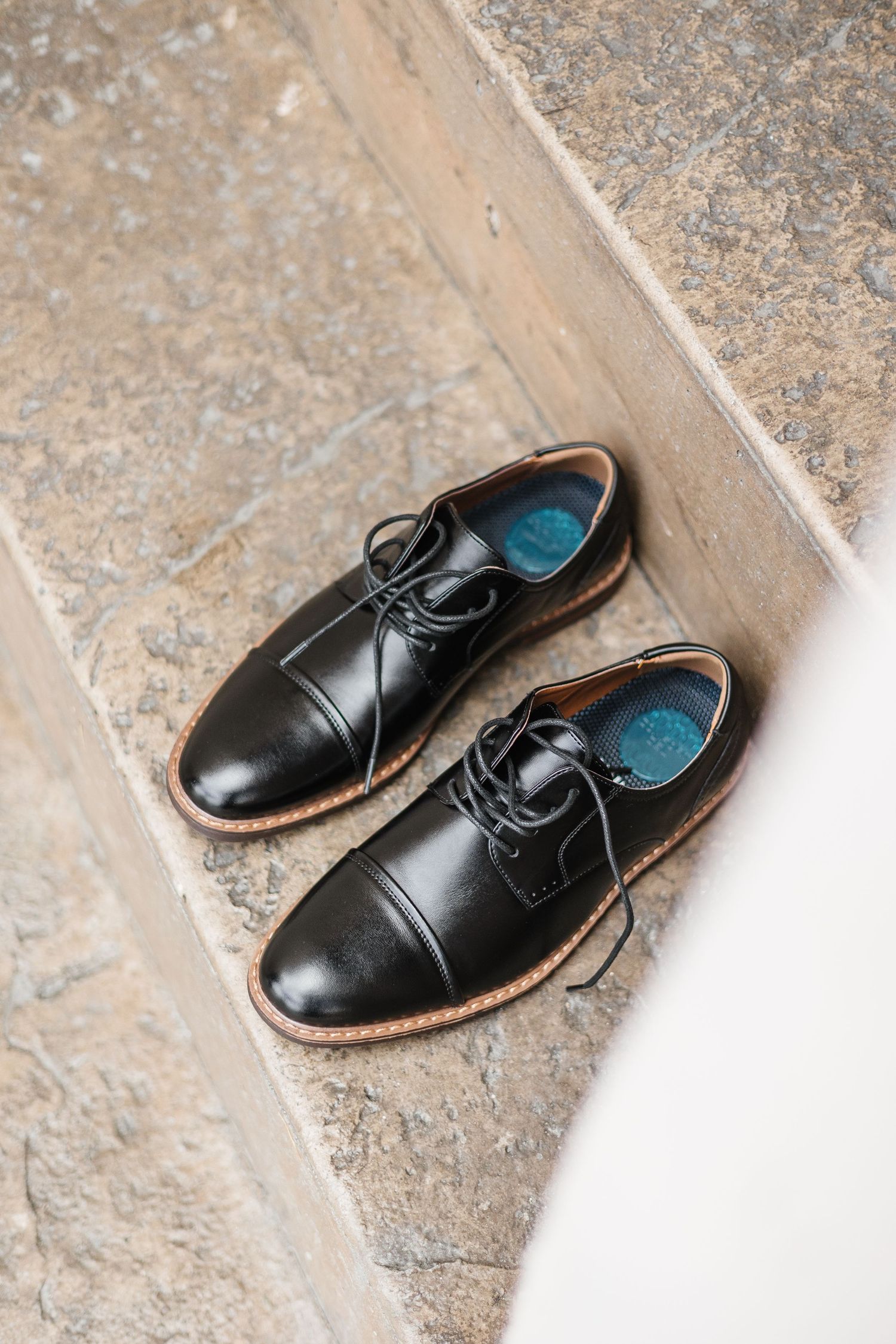A pair of classic black leather dress shoes with laces displayed on a textured stone floor.