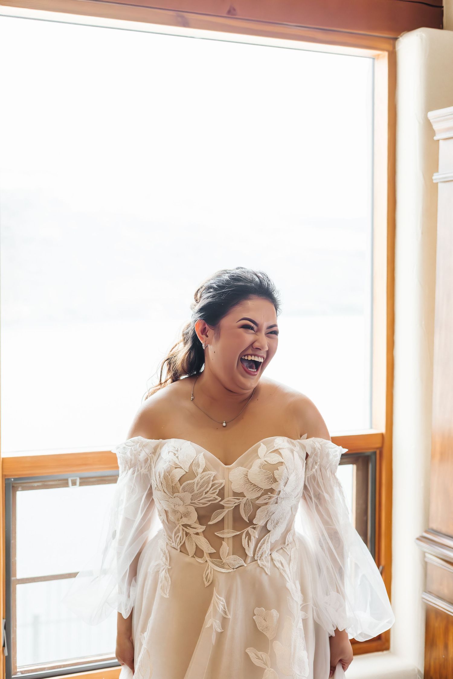A bride in an off-shoulder wedding gown laughs joyfully during her preparation.