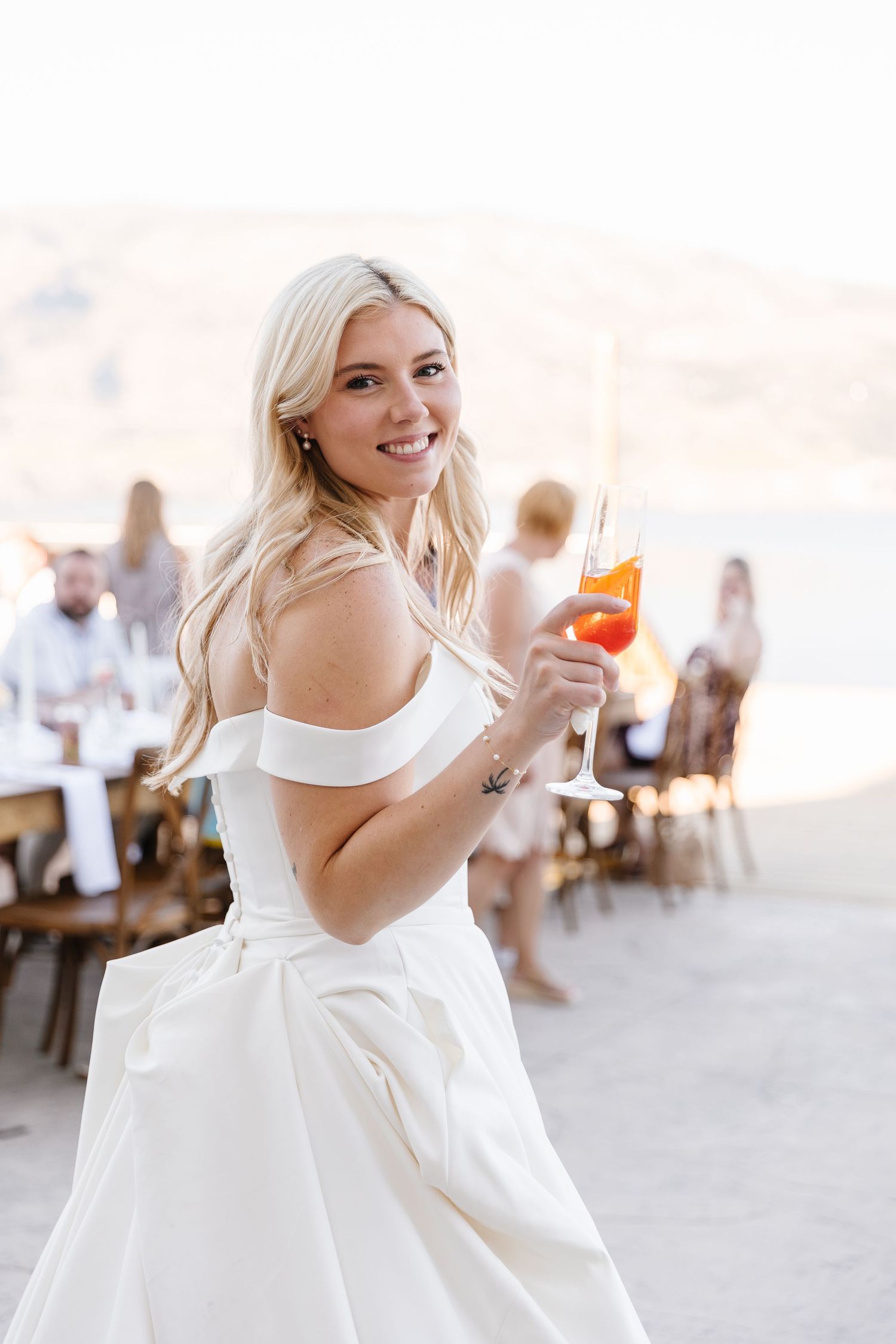 A bride in a white dress laughs joyfully while holding a champagne glass at an outdoor reception.