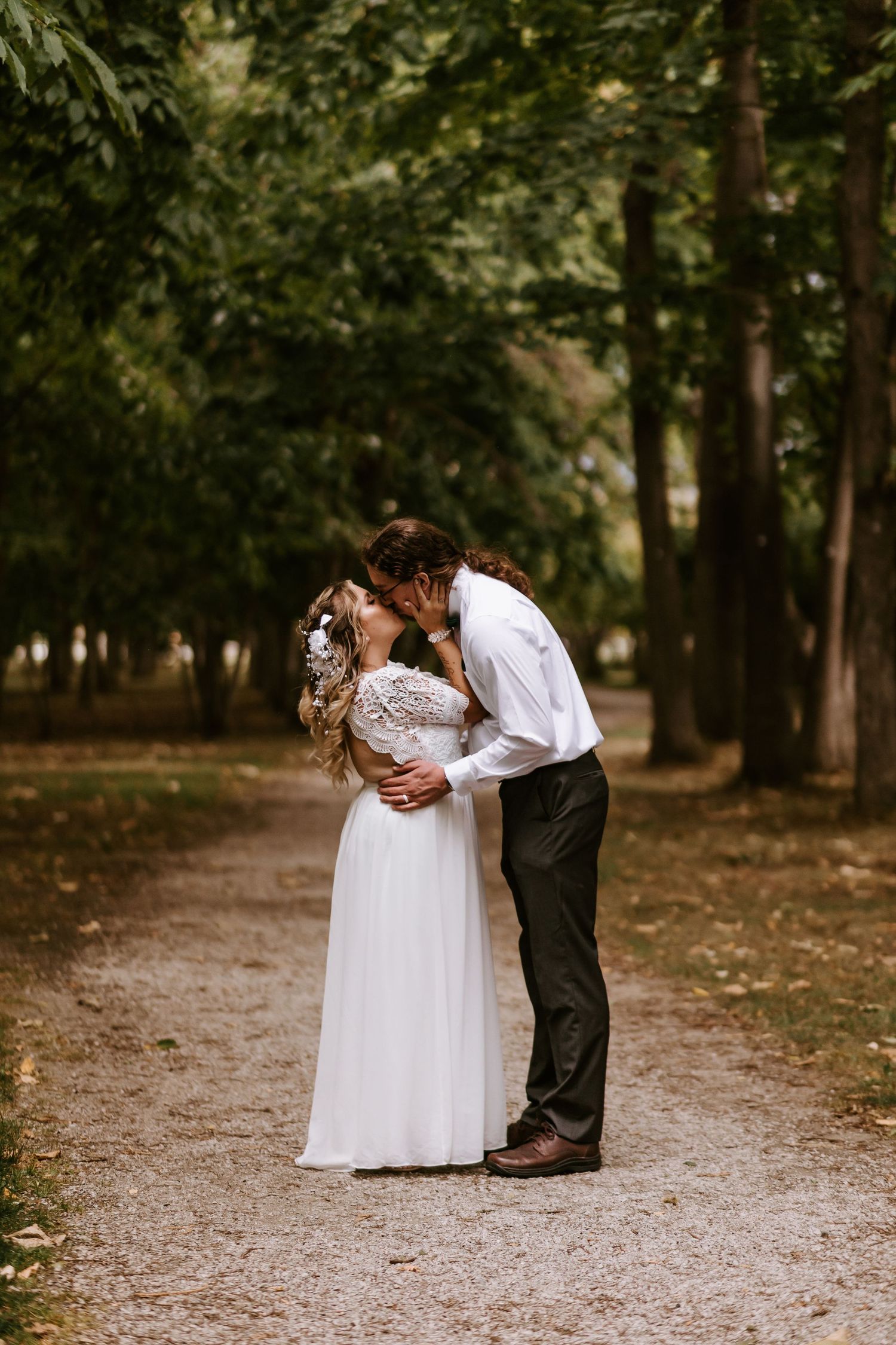 A romantic couple in wedding attire embrace on a tree-lined gravel path in a park setting.