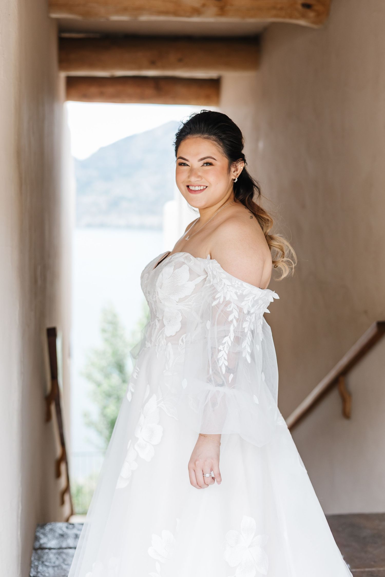 A bride in an off-shoulder white lace wedding gown with flowing sleeves poses in a rustic hallway with mountain views.