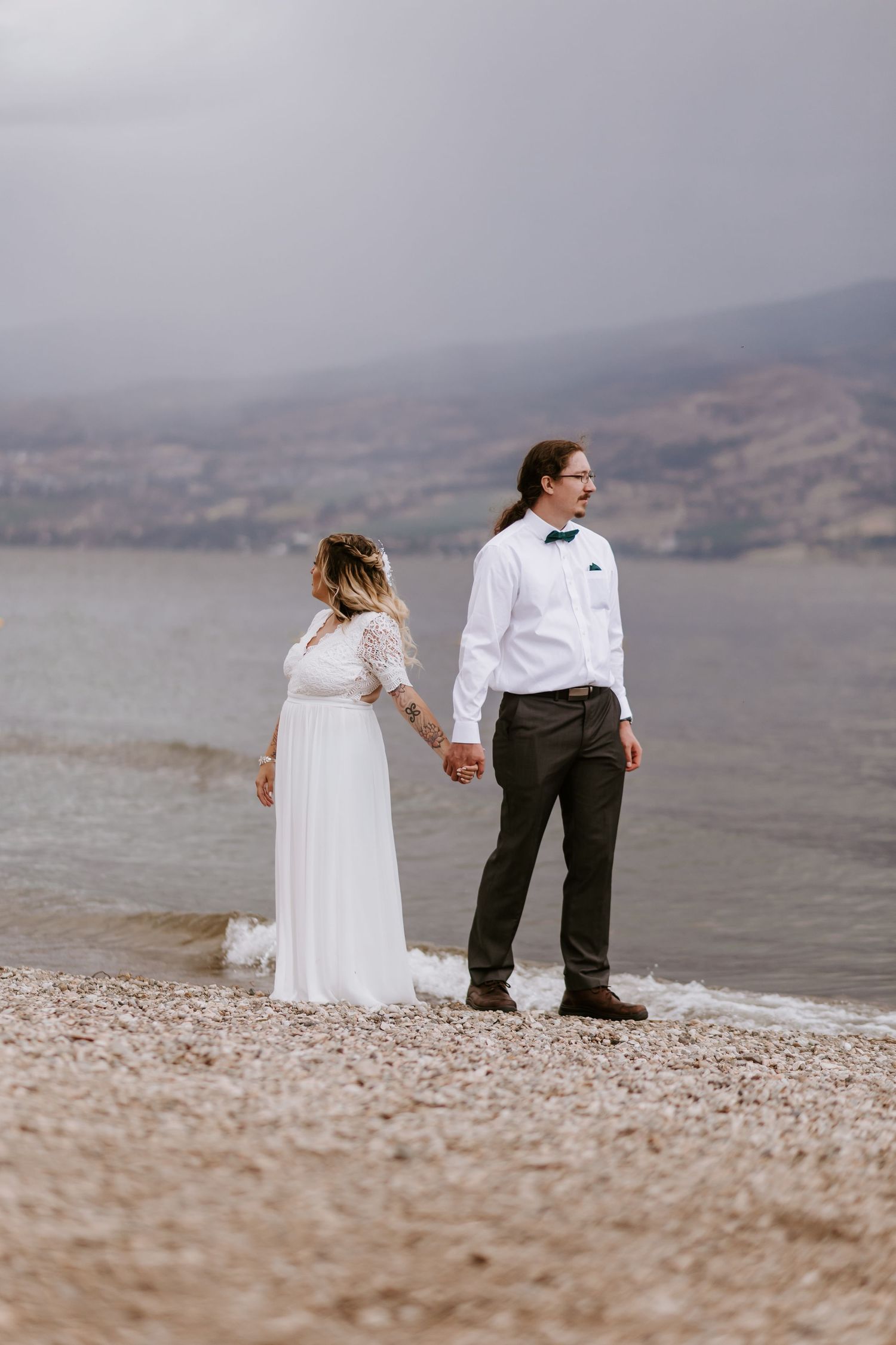 A couple stands together holding hands on a rocky beach shore with misty mountains in the background.