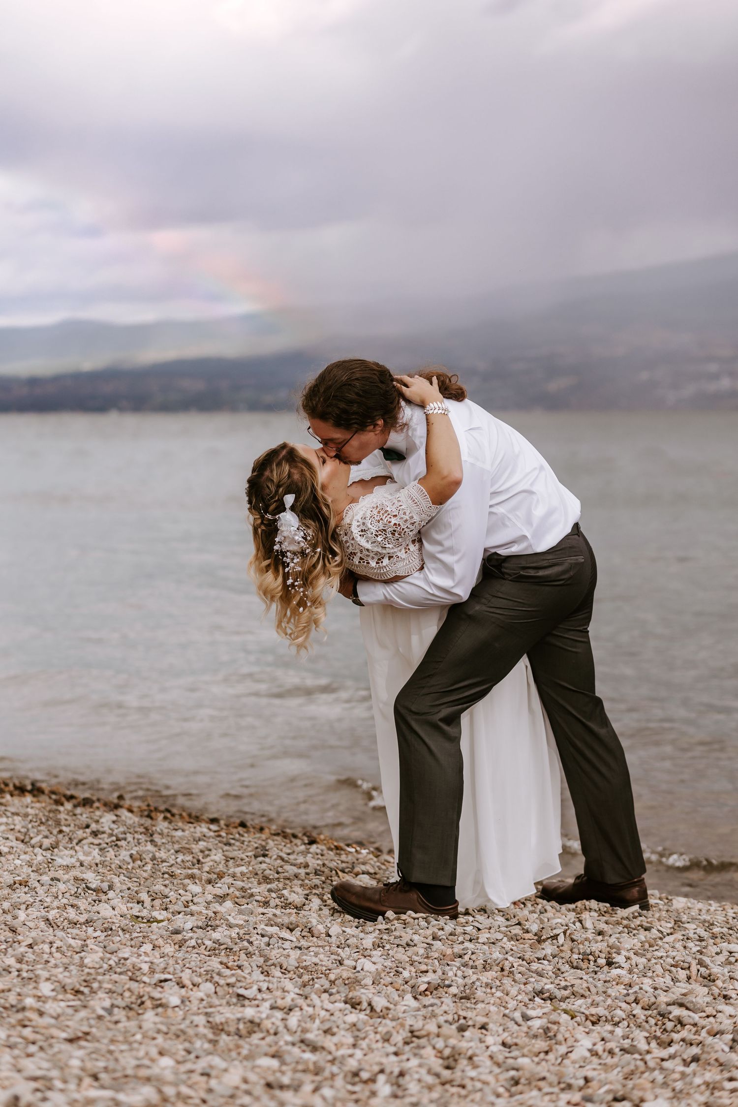 A romantic couple embraces on a pebble beach shoreline with mountains and moody skies in the background.