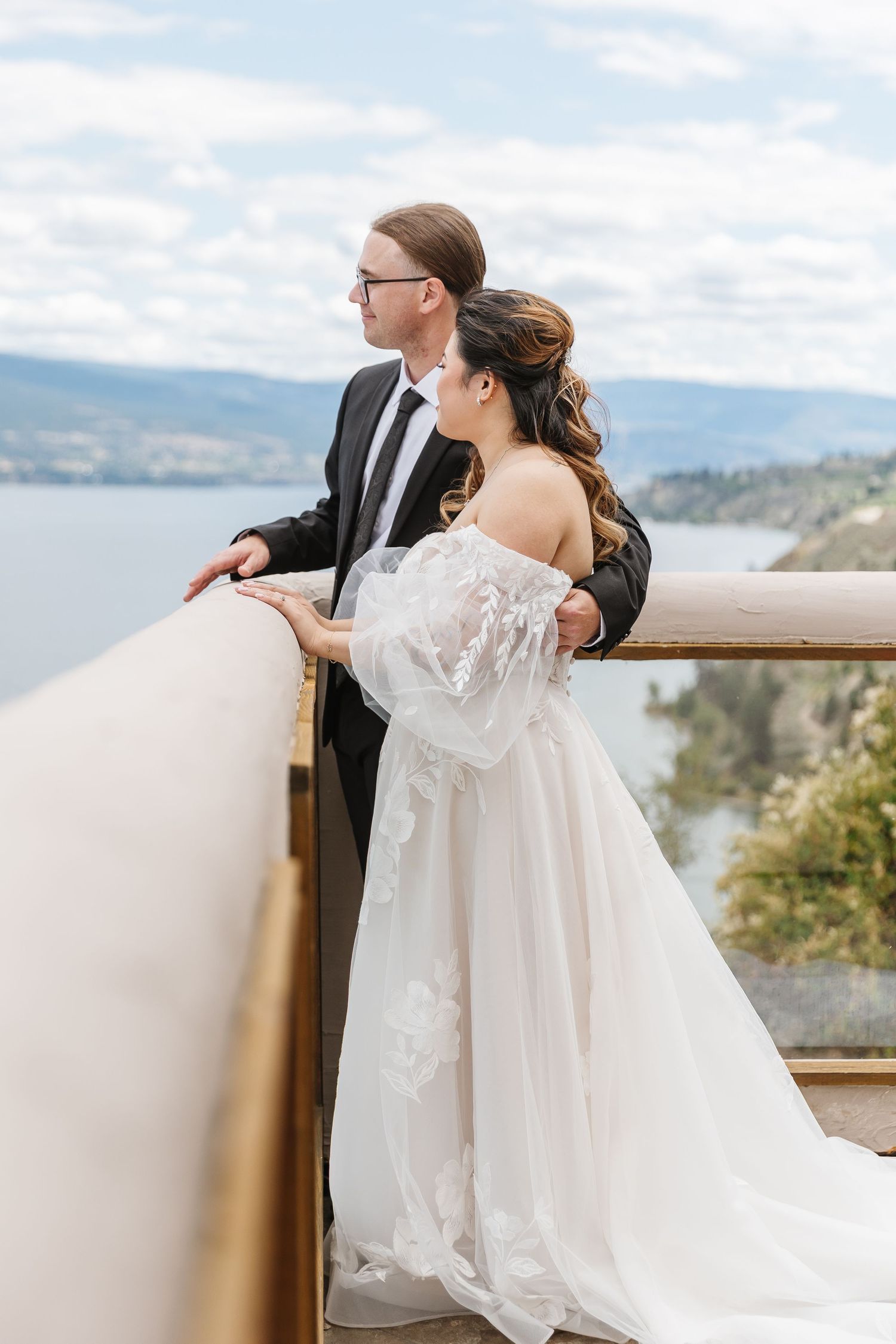 A couple in wedding attire gazes at a scenic mountain landscape from a balcony overlooking a lake.