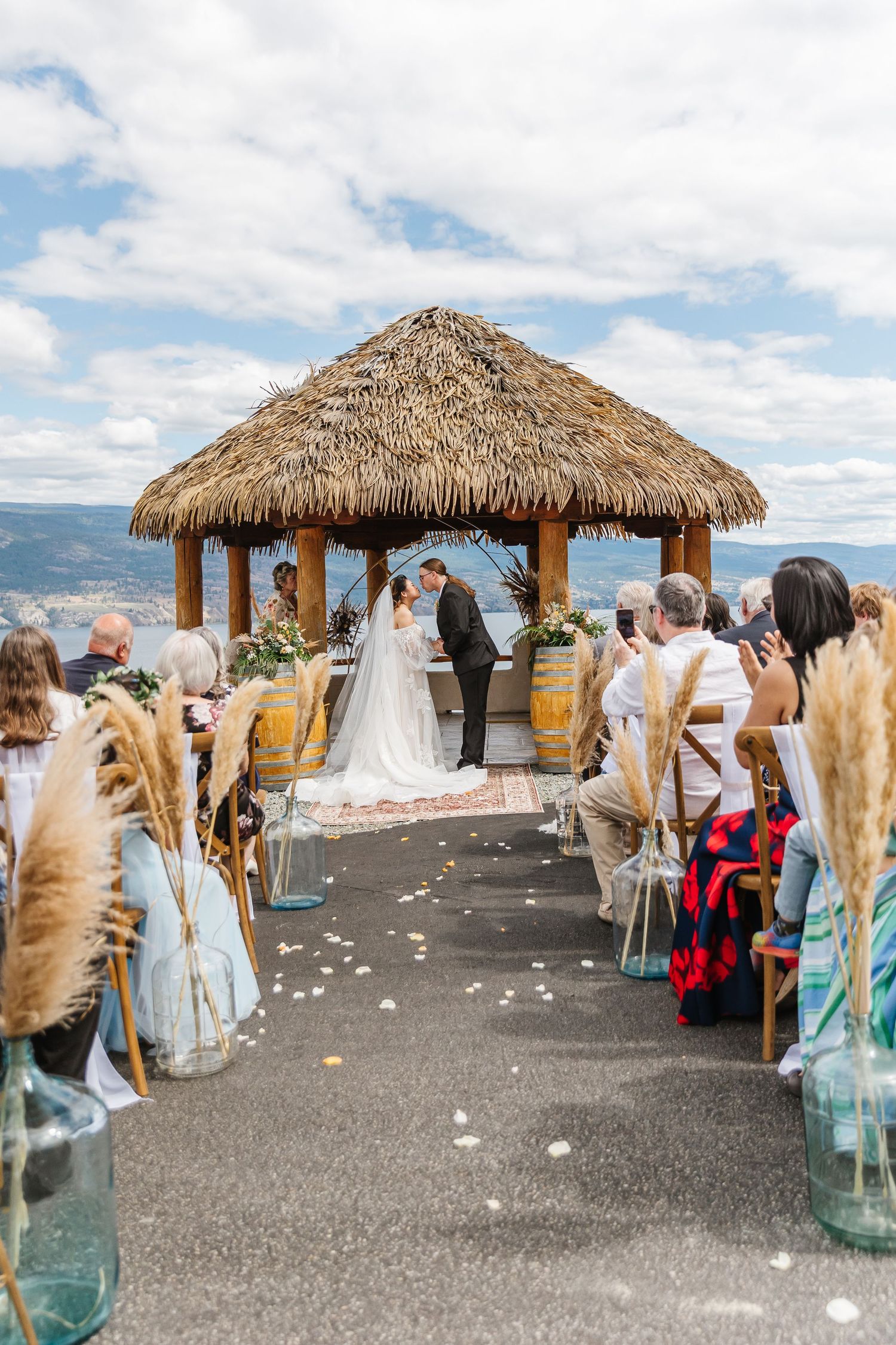 Outdoor wedding ceremony under a thatched-roof gazebo overlooking mountains with guests seated on both sides.