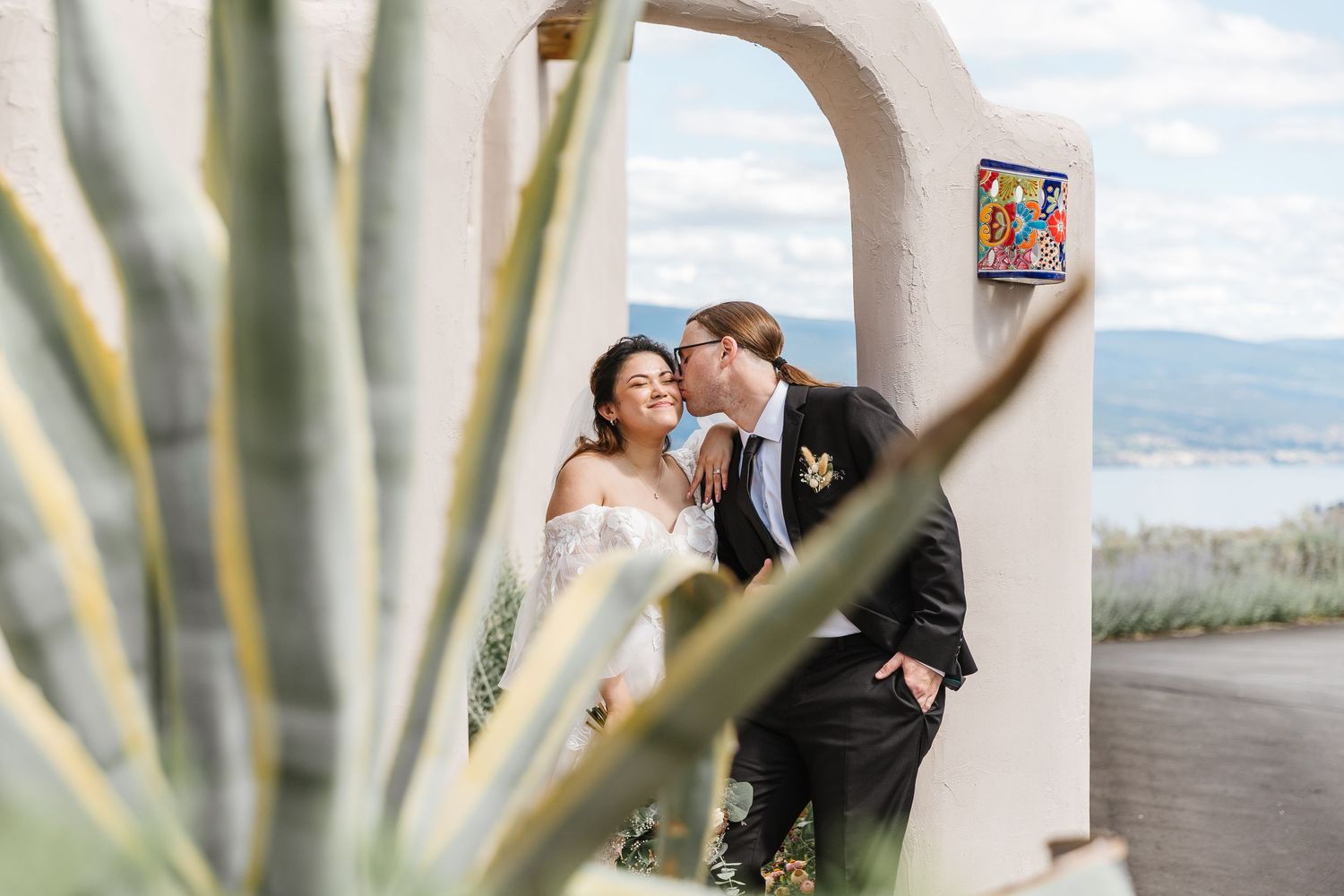 A couple shares a romantic moment under a white archway by the beach, framed by tropical agave plants in the foreground.