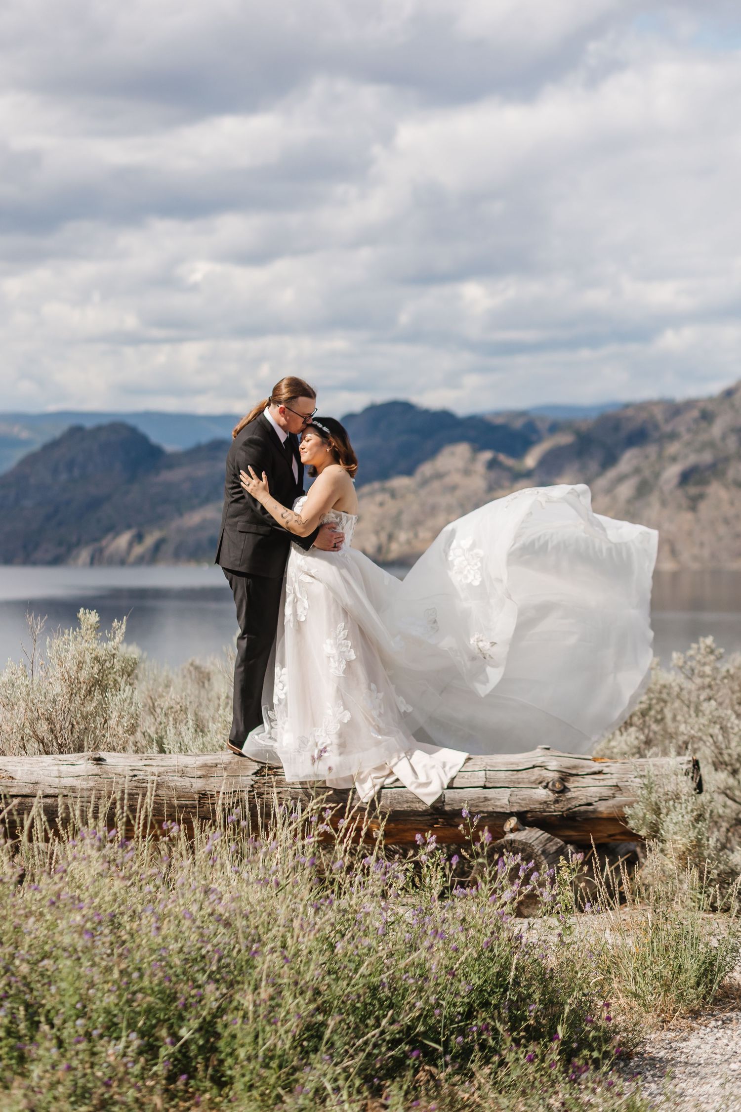 Romantic wedding couple embraces on a mountain overlook with flowing veil and dramatic landscape.