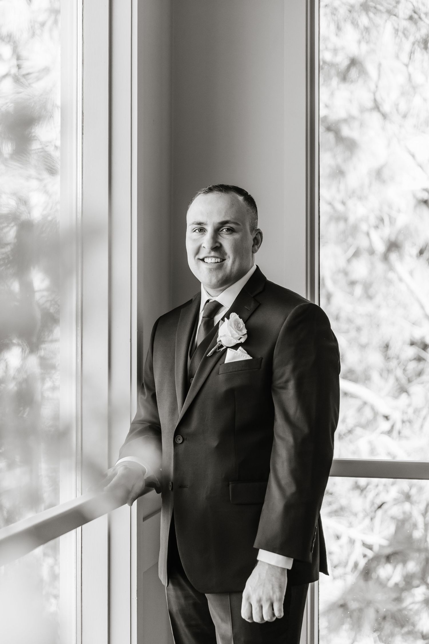 Black and white portrait of a groom in a formal suit smiling by a bright window.