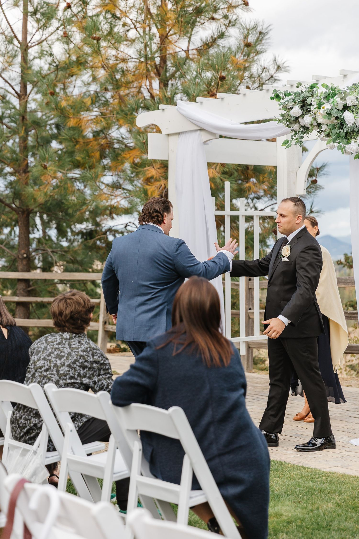 Wedding guests and attendees gather at an outdoor ceremony with a white draped arch and folding chairs on the lawn.