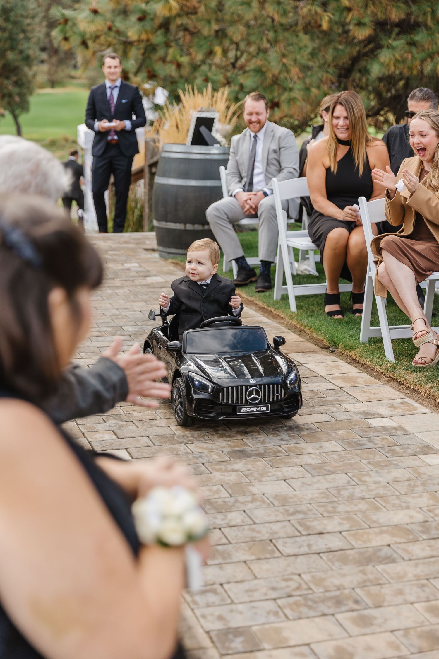 Children drive miniature Mercedes toy cars down a stone pathway at an outdoor wedding ceremony.