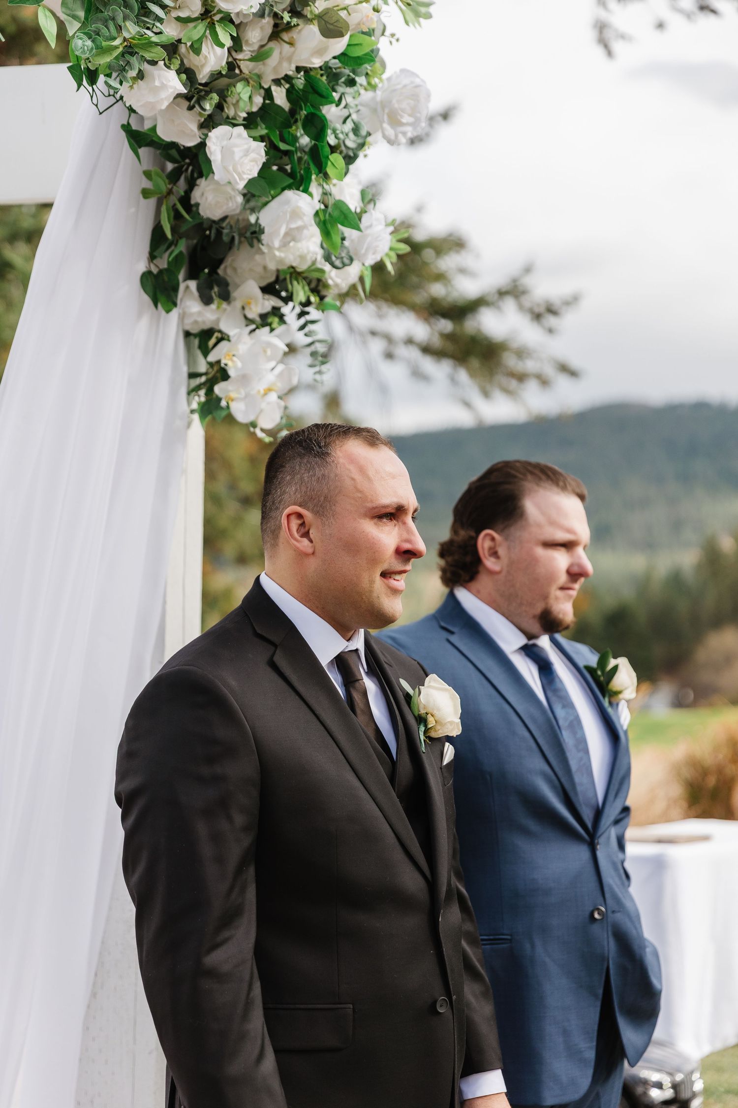 Two groomsmen stand at attention under a floral-decorated white arch with mountains visible in the background.