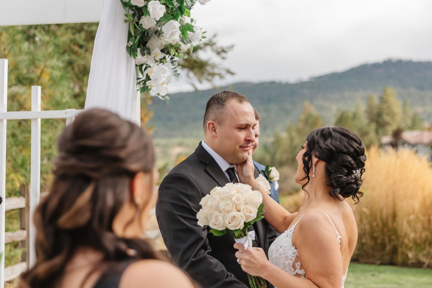 Wedding guests sit in white chairs at an outdoor ceremony venue with mountain views and floral decorations.