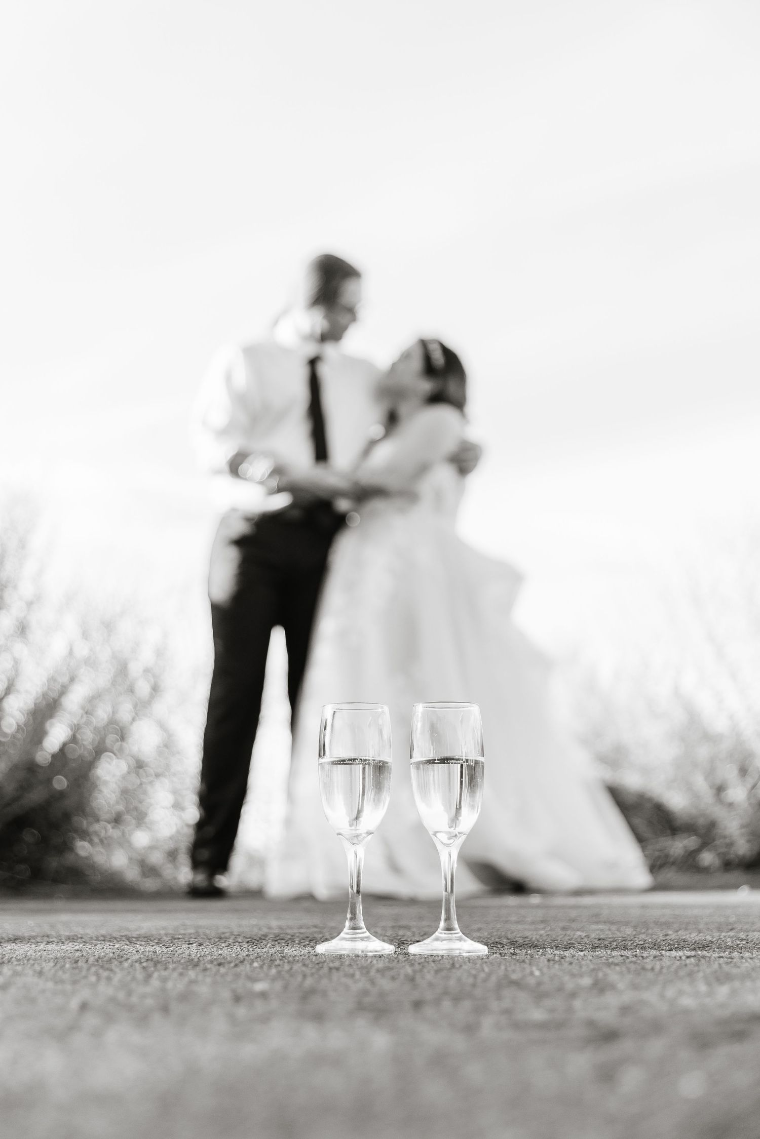 Two champagne glasses in foreground with blurred newlyweds in background, black and white photography.