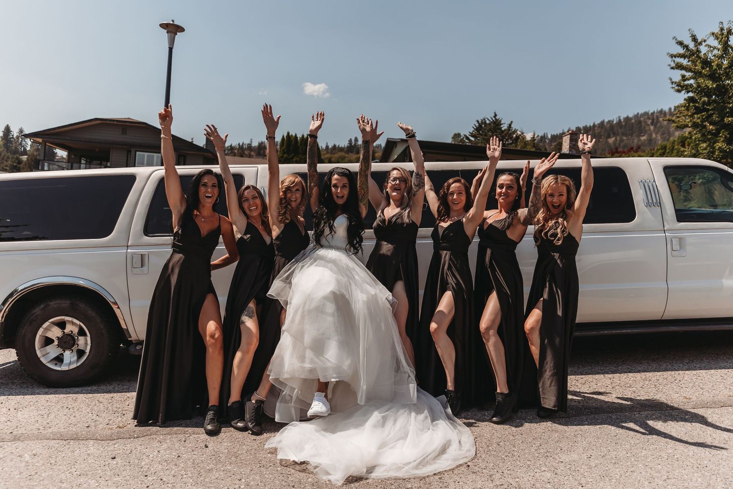Wedding party poses in front of white limousine with bridesmaids in black dresses and bride in flowing white gown.