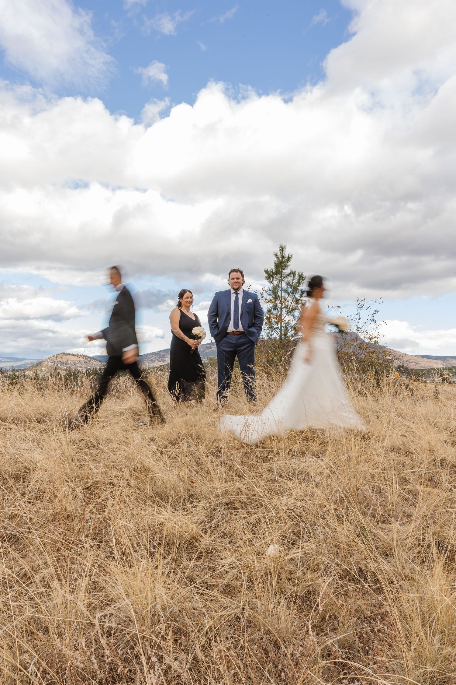 Wedding party stands in golden grass field with mountains and cloudy sky in background during outdoor celebration.
