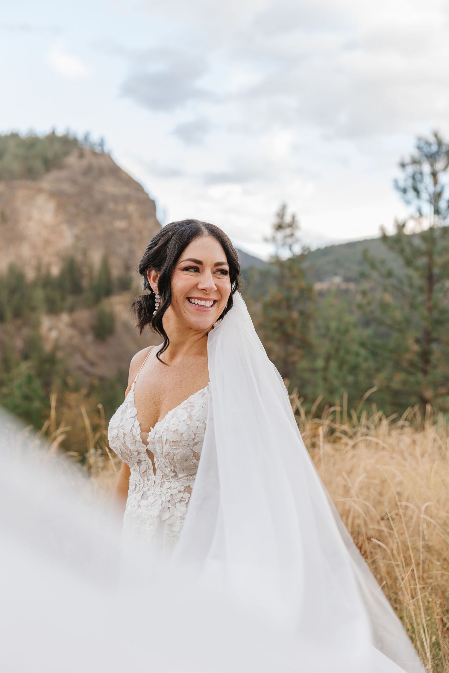 A bride in a lace wedding dress and veil poses in a field of tall golden grass with mountains in the background.