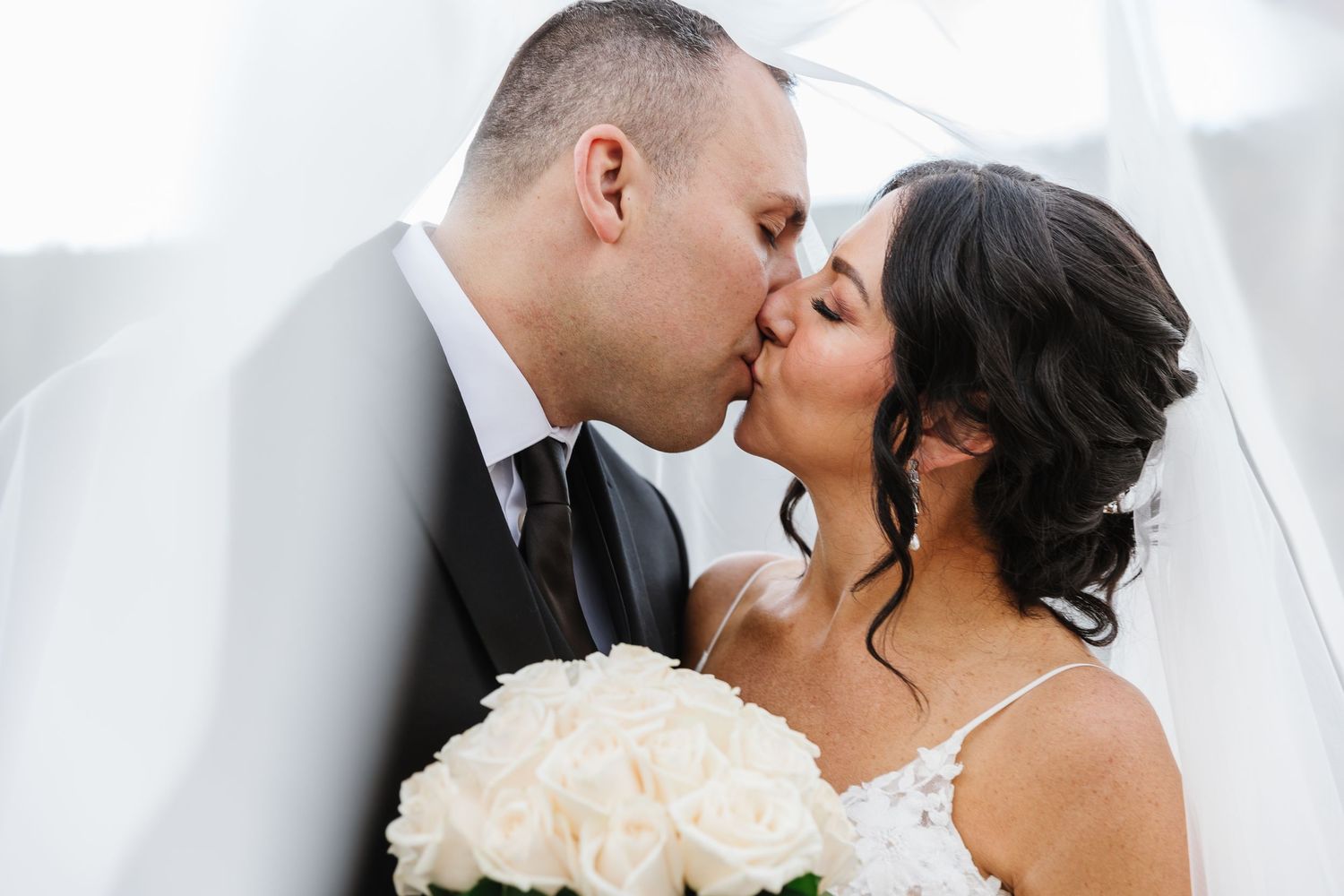 A romantic wedding couple shares a tender kiss while holding a white rose bouquet.