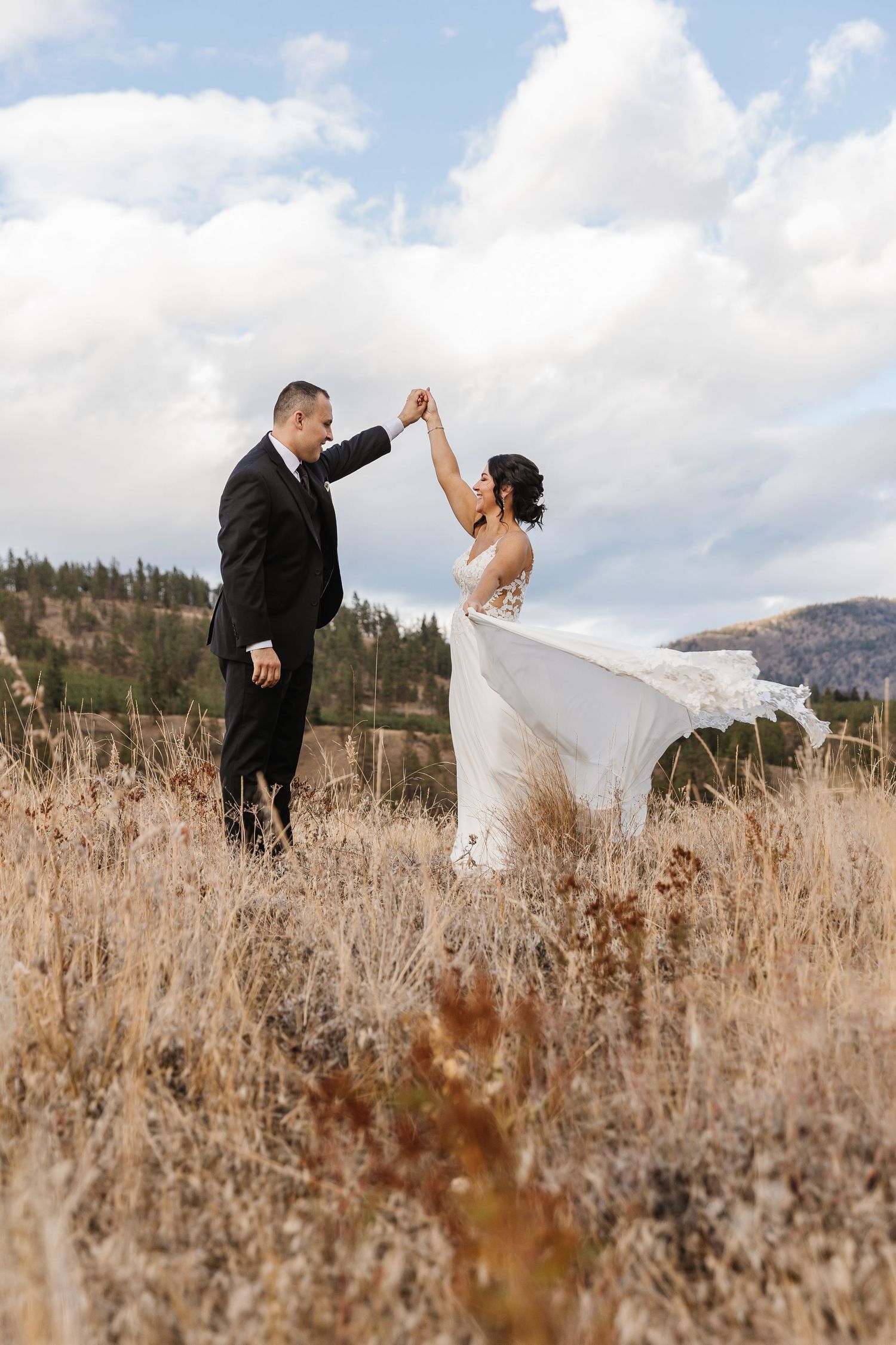 A couple in wedding attire dance together in a field of tall grass with mountains and cloudy skies in the background.