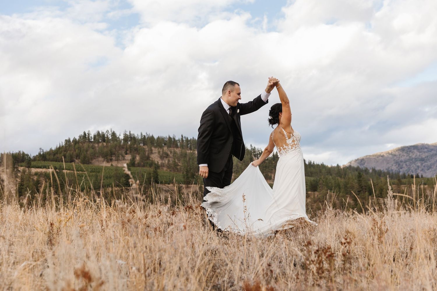 A couple in wedding attire stands in a golden field with mountains and evergreen trees in the background.