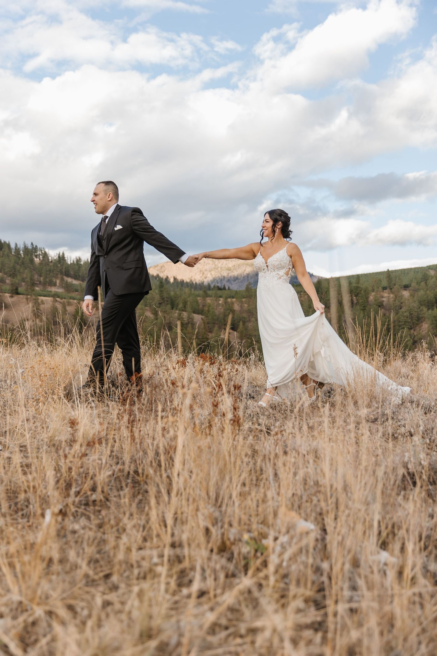 A couple playfully poses in a golden field with mountains and forest in the background during their wedding shoot.