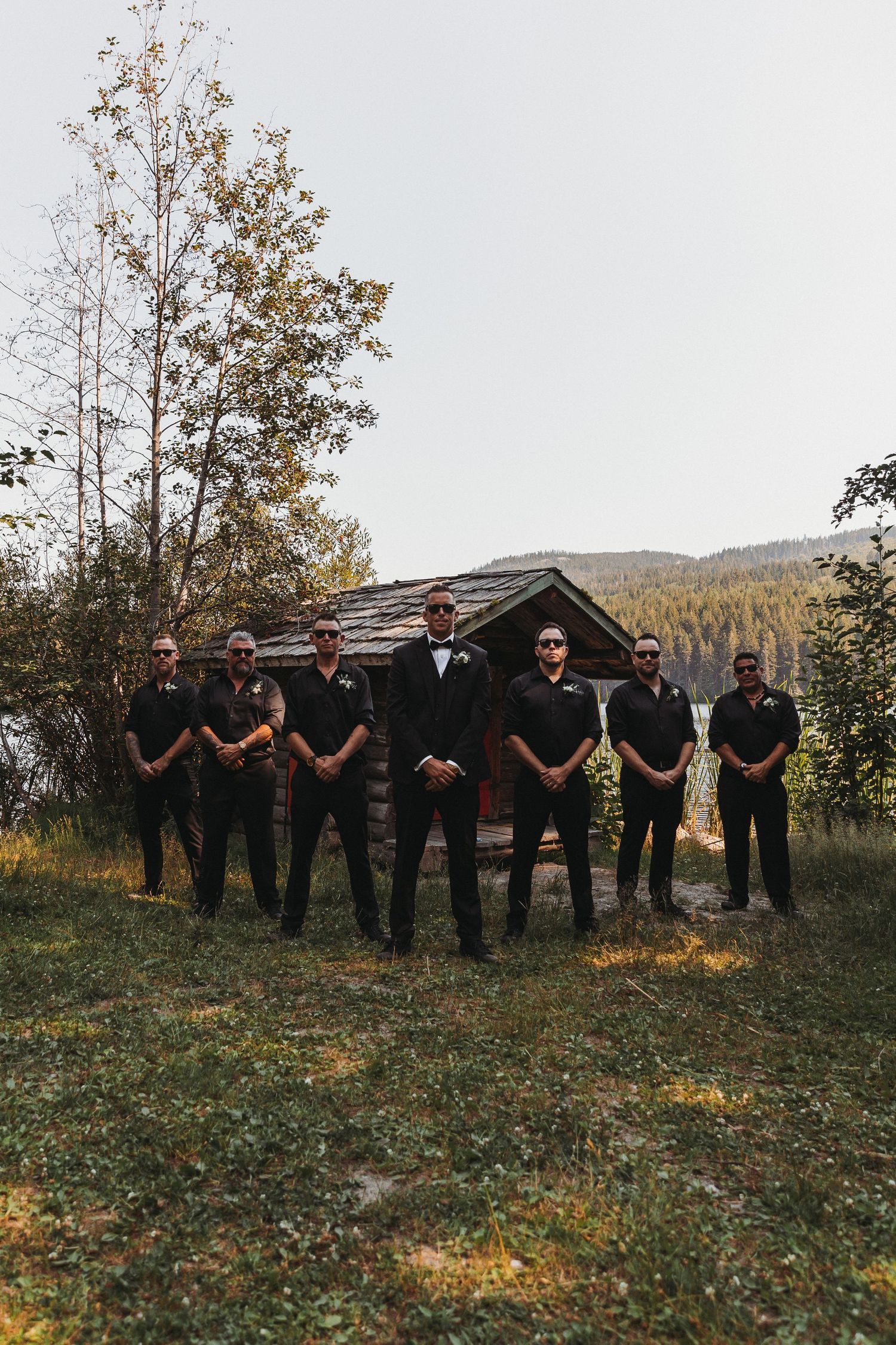 Wedding party in black attire poses on a grassy hillside with autumn trees in the background.