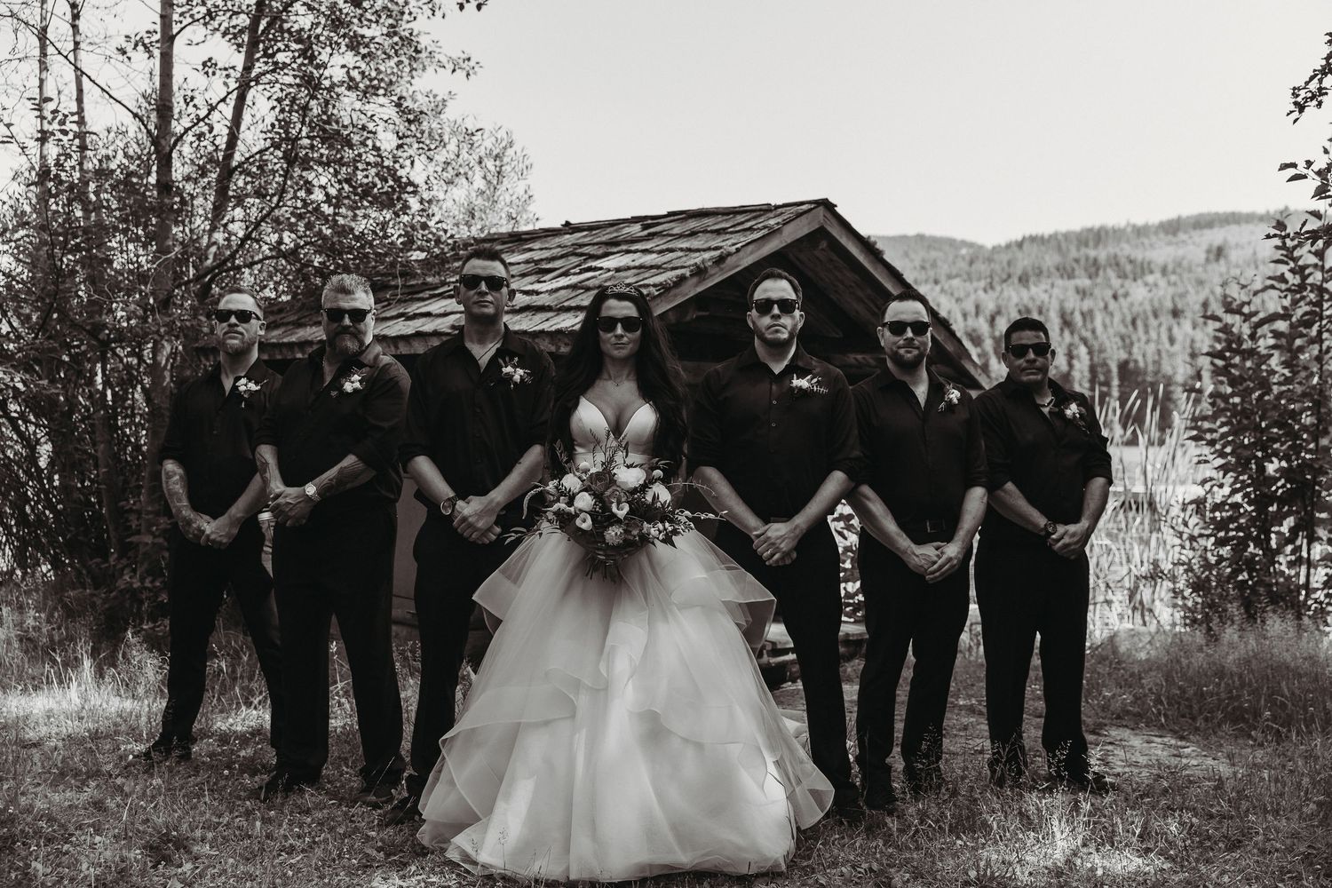 Wedding party in black attire poses together with bride in white dress against natural outdoor backdrop.