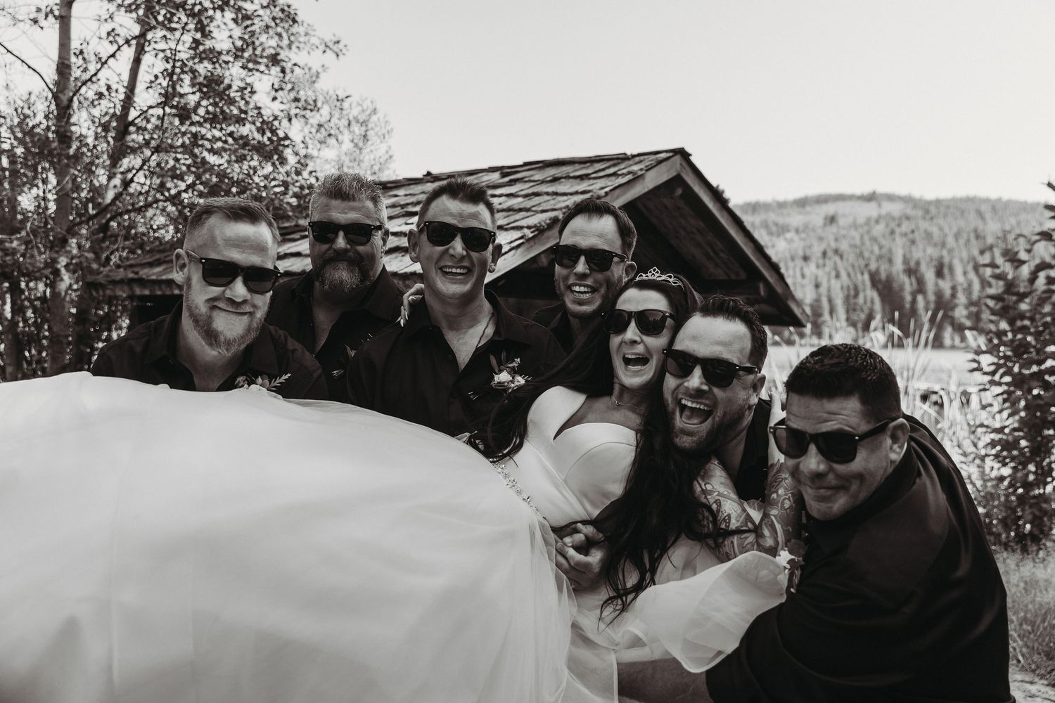 A black and white photo of a group wearing sunglasses gathered together outdoors with a rustic cabin in the background.