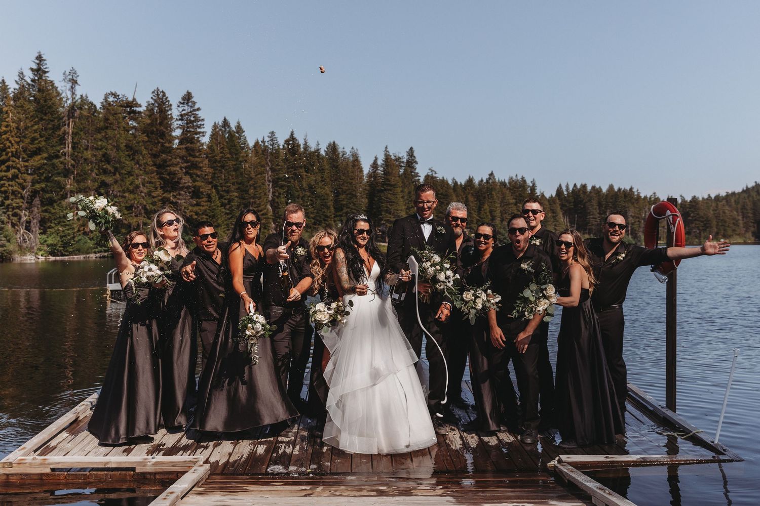 Wedding party poses together on a wooden dock by a scenic lake with evergreen trees in the background.