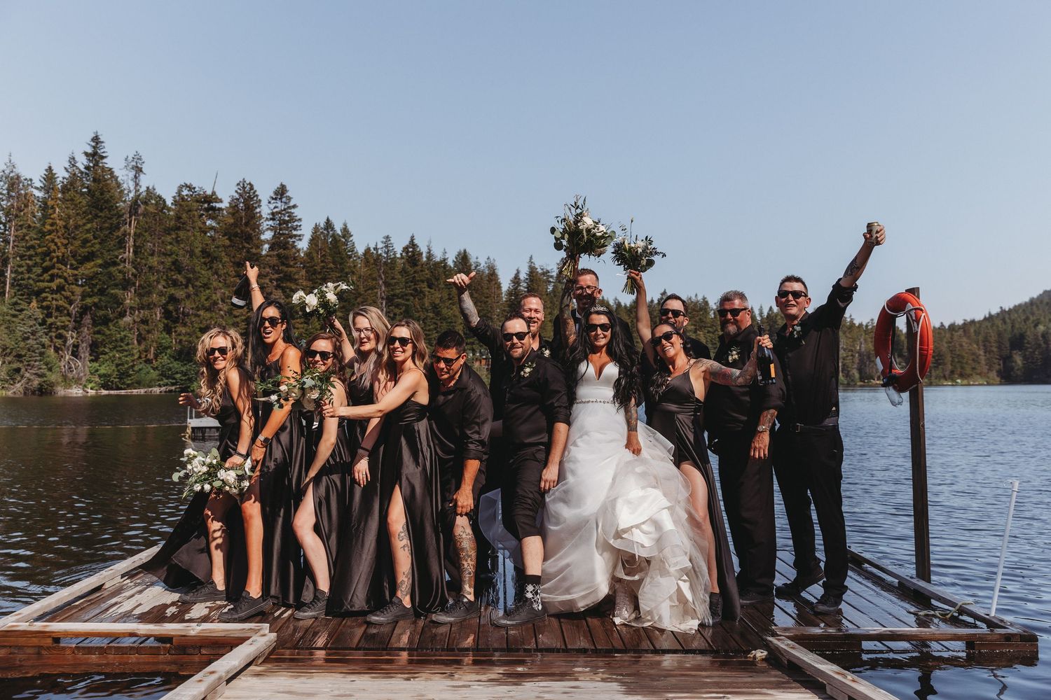 Wedding celebration group poses joyfully on a wooden dock by a scenic mountain lake.