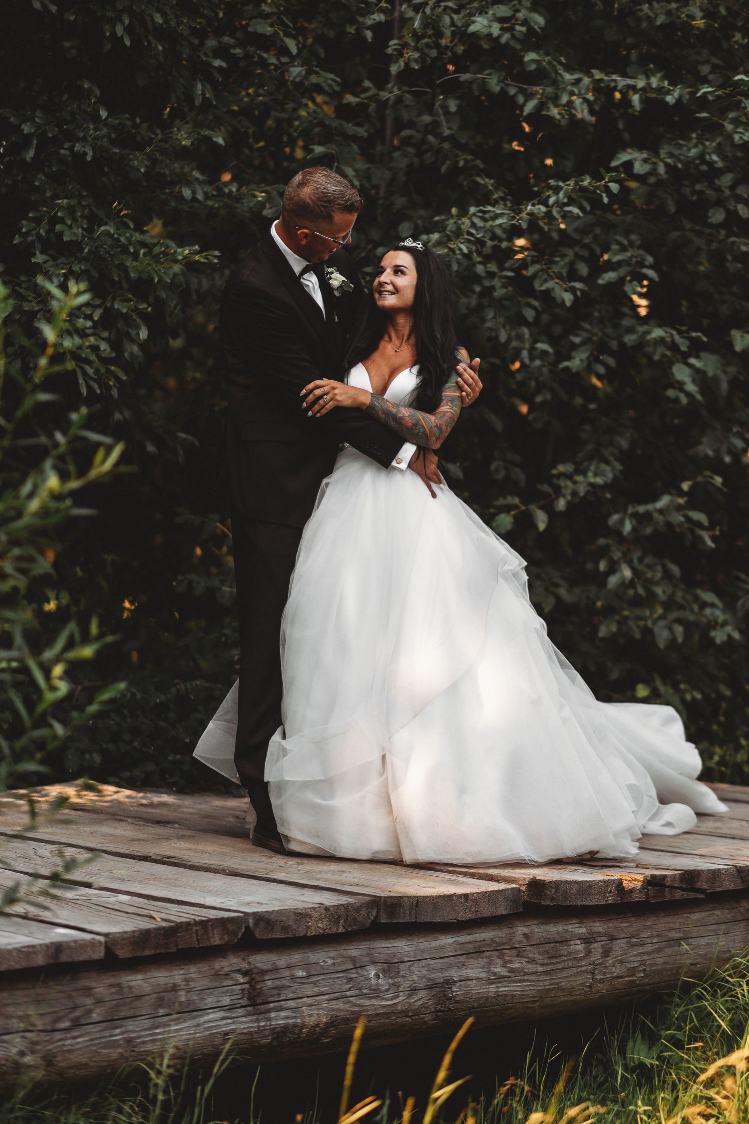 A romantic wedding couple stands together on a rustic wooden dock surrounded by lush greenery.