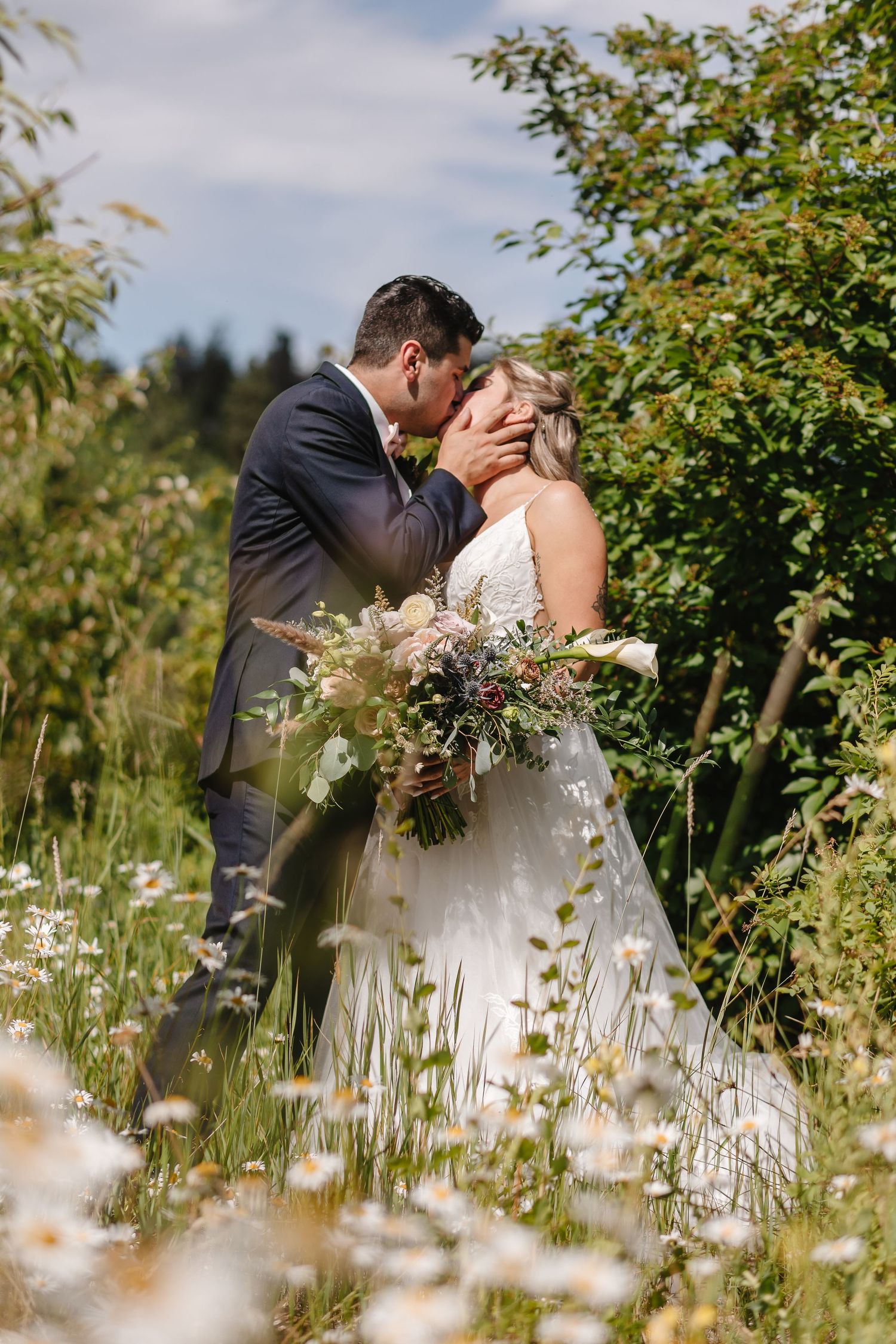 A couple shares a romantic kiss in a meadow filled with white wildflowers on their wedding day.