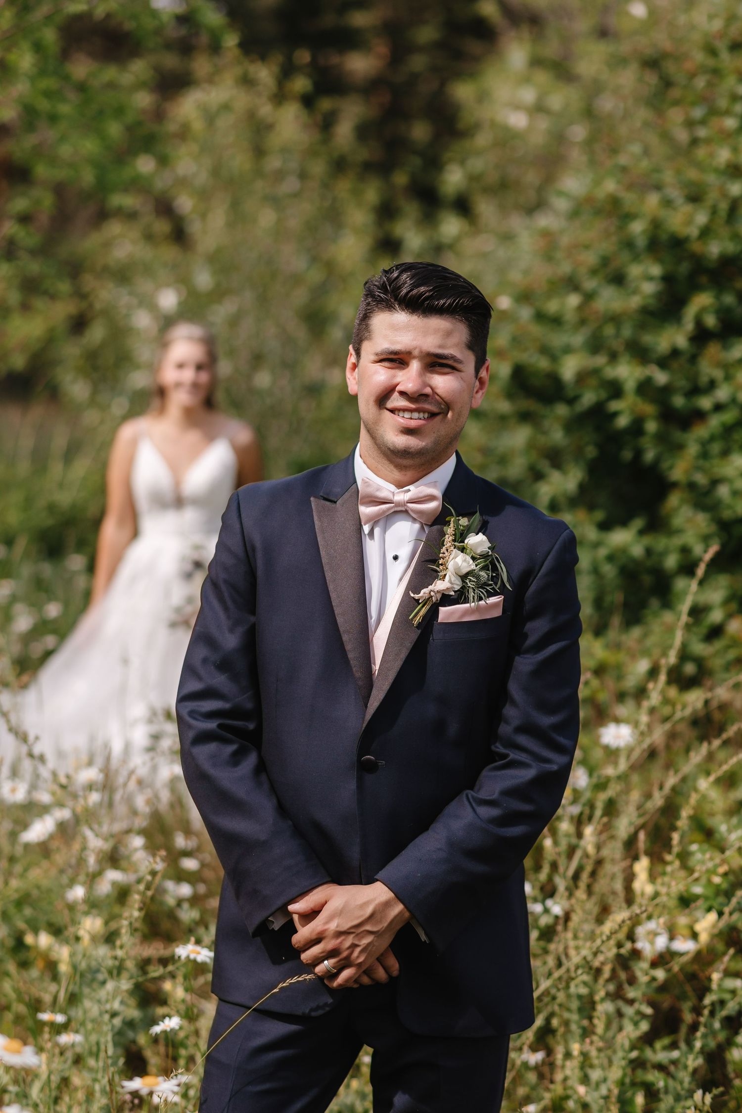 A groom in a navy suit stands in a sunny garden setting during a wedding photoshoot.