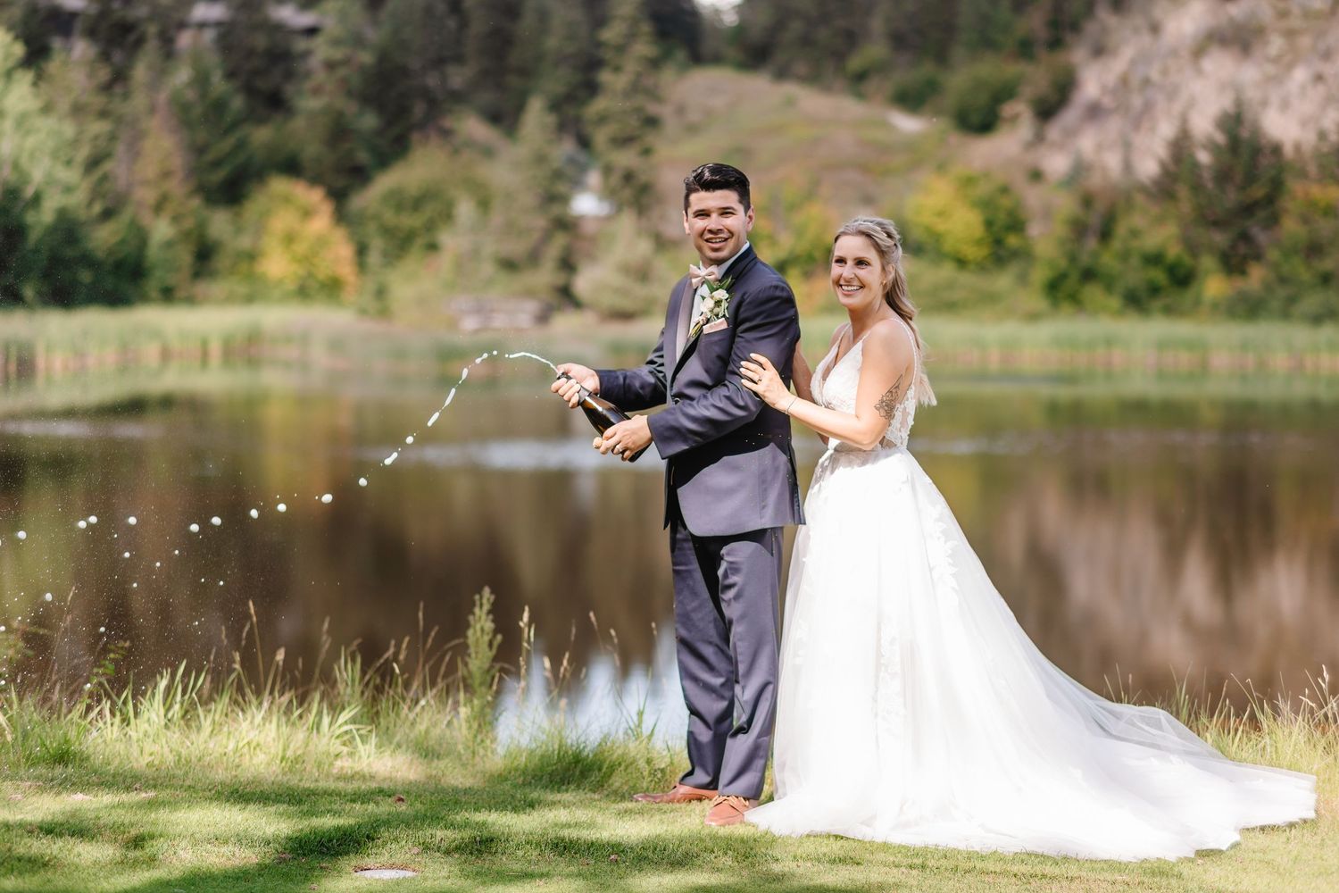 A couple in wedding attire stands by a serene lake surrounded by evergreen trees in a mountain setting.