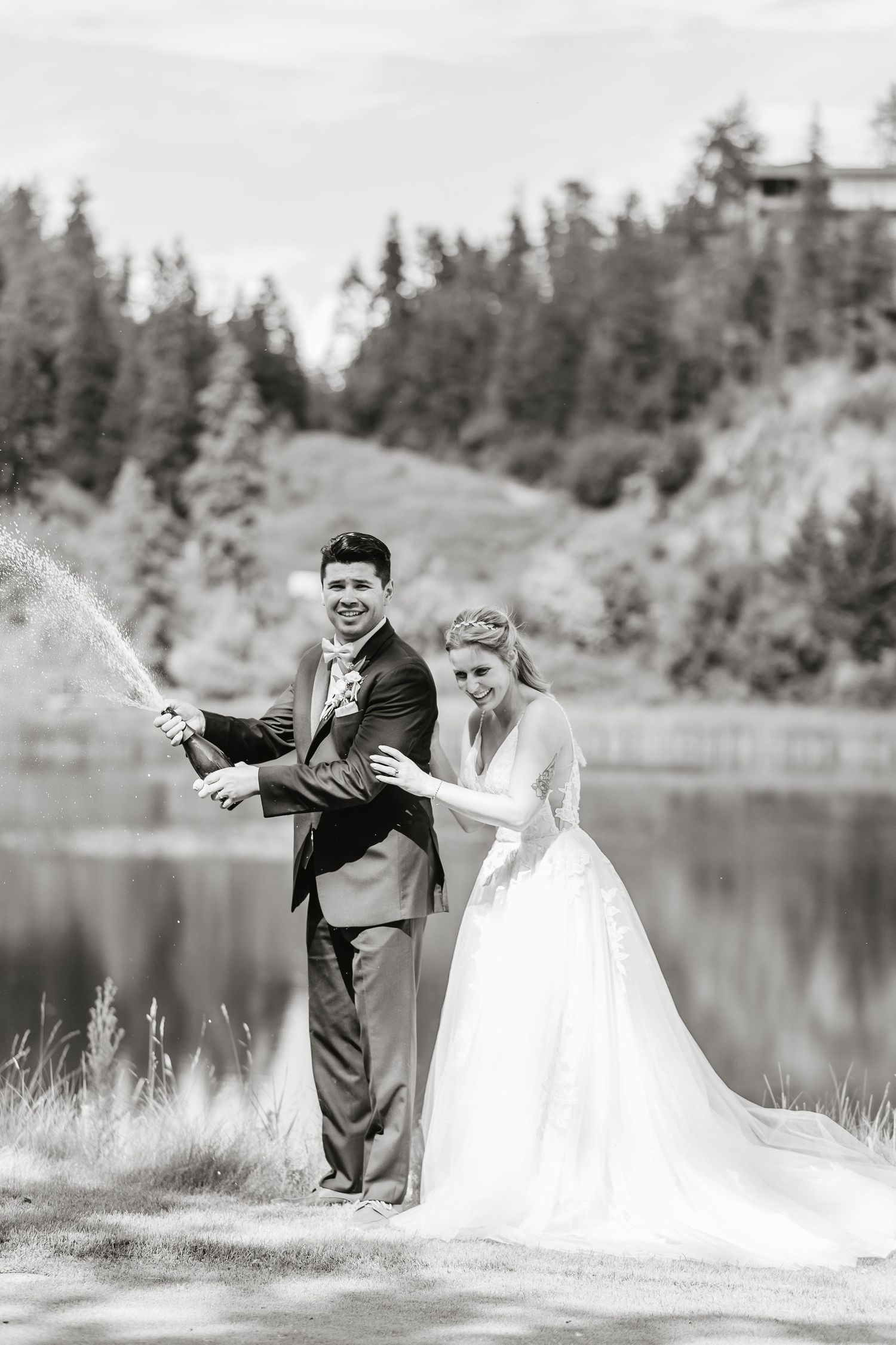 A playful wedding couple poses for a sequence of photos by a serene lake surrounded by evergreen trees.