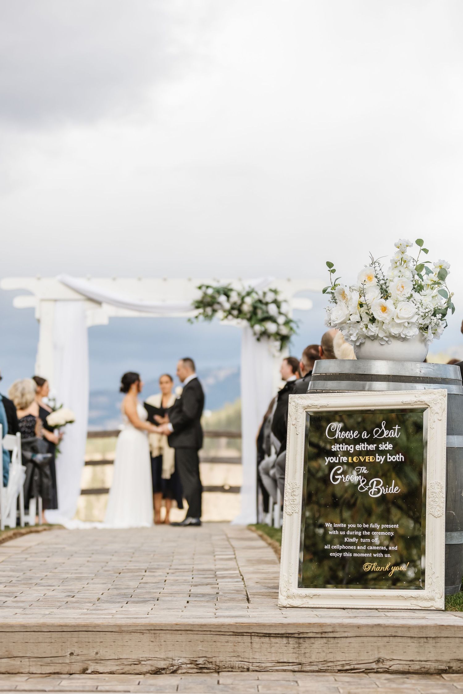 Wedding ceremony setup with chalkboard signs and white floral arrangements against a mountain backdrop.