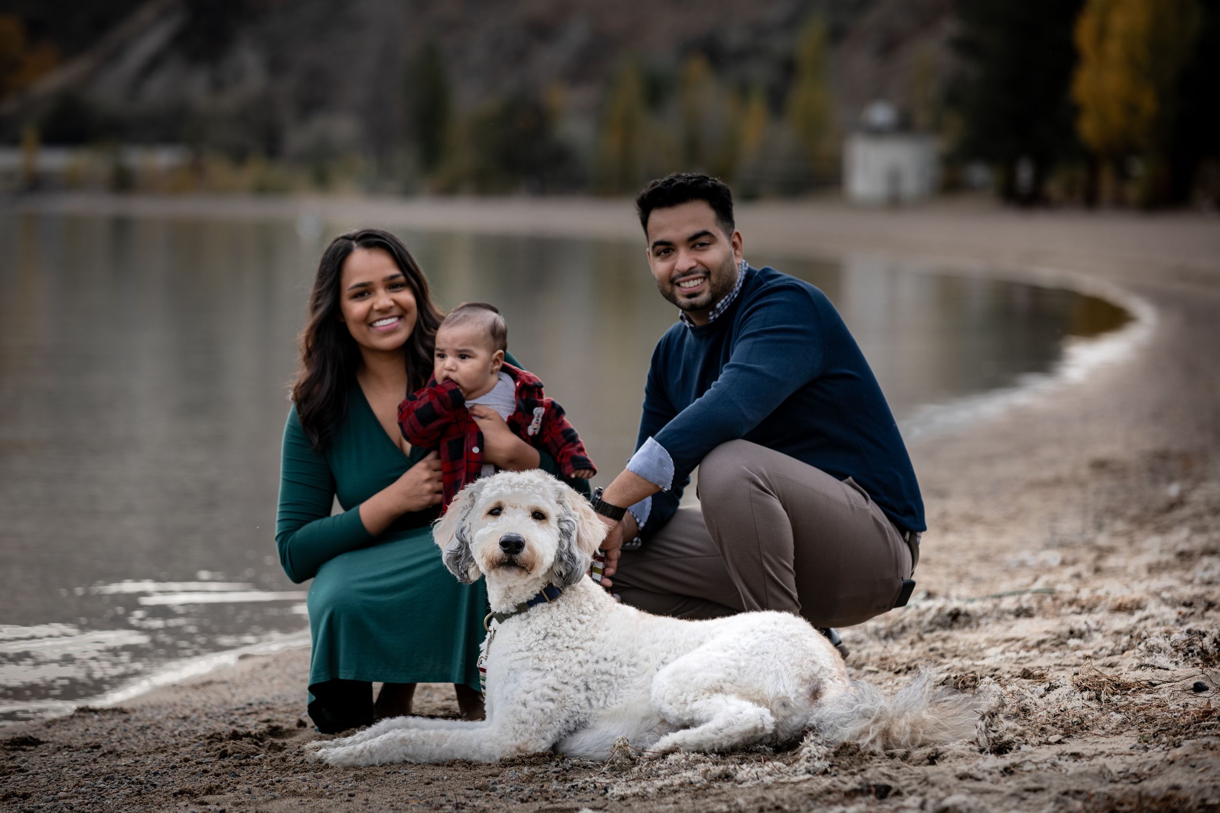 Happy family with baby and white dog sitting together on rocky lakeside beach.