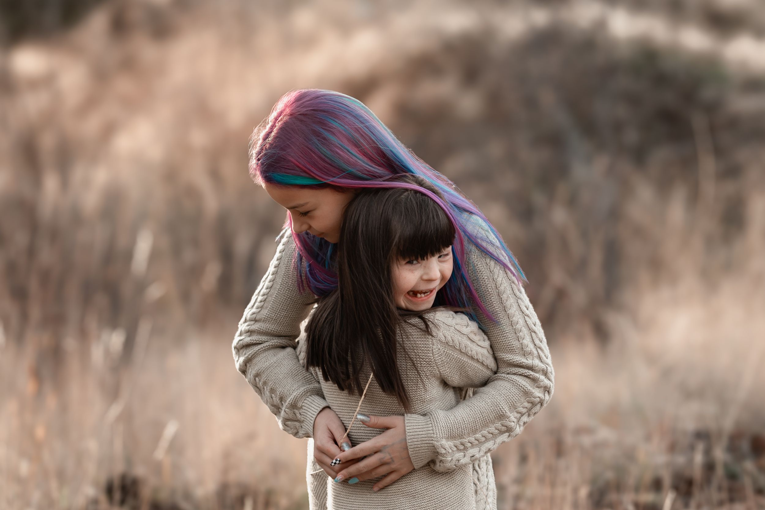 Woman with colorful hair tenderly embracing young girl in dry grass field outdoors.