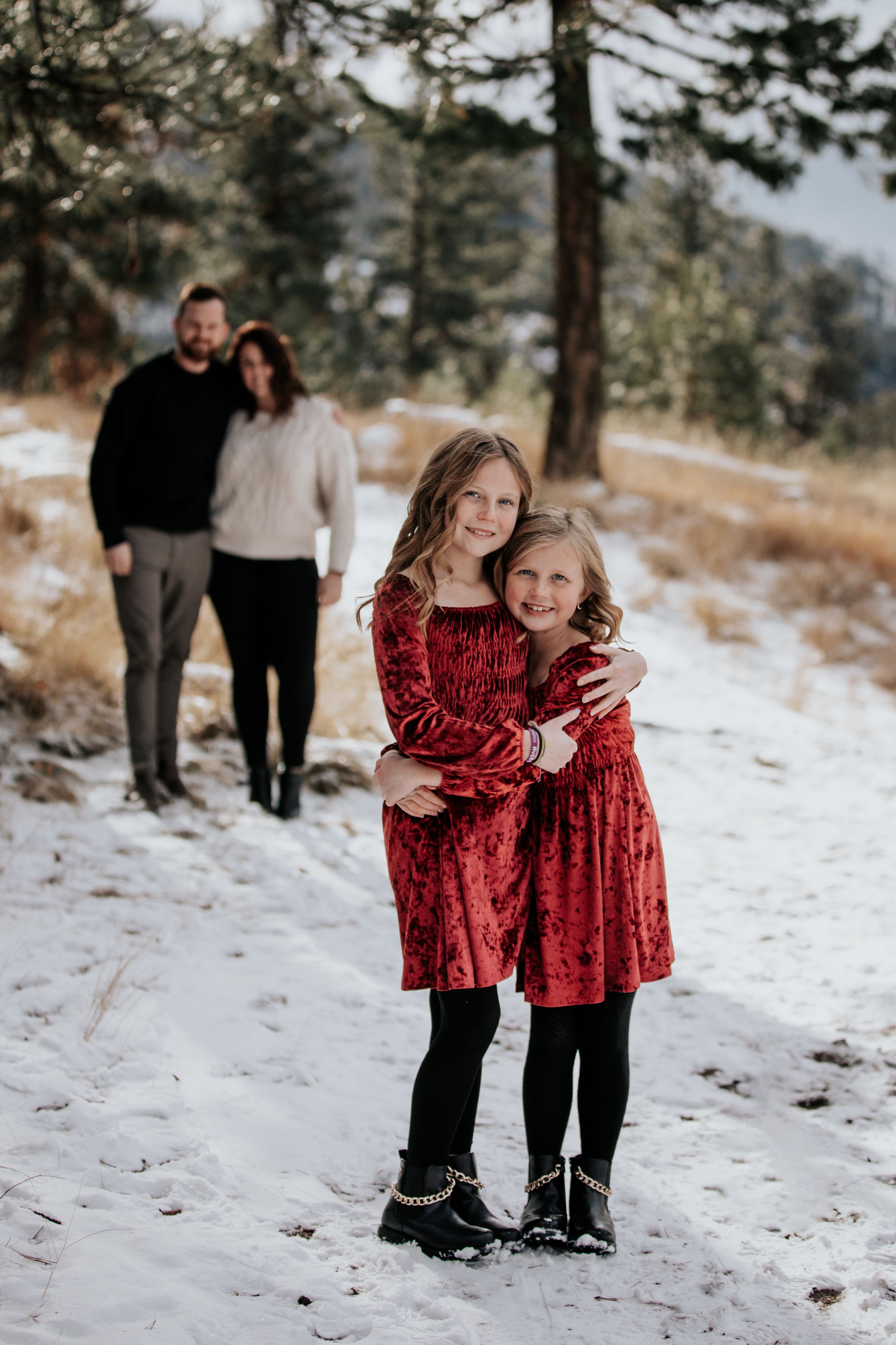 Two sisters in red dresses hugging on snowy mountain path with parents behind them.