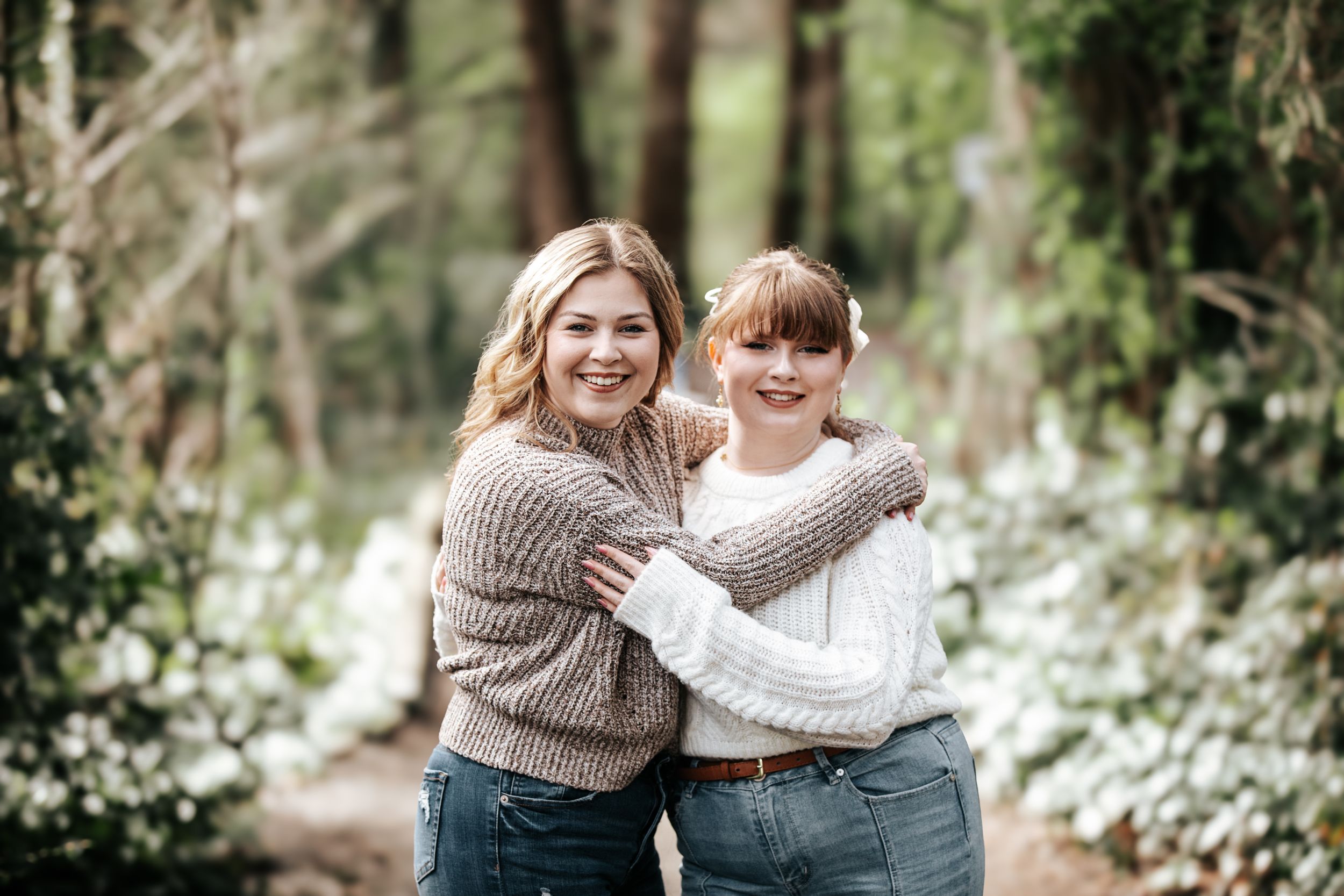 Smiling mother and daughter hugging in white flower garden wearing matching brown sweaters.