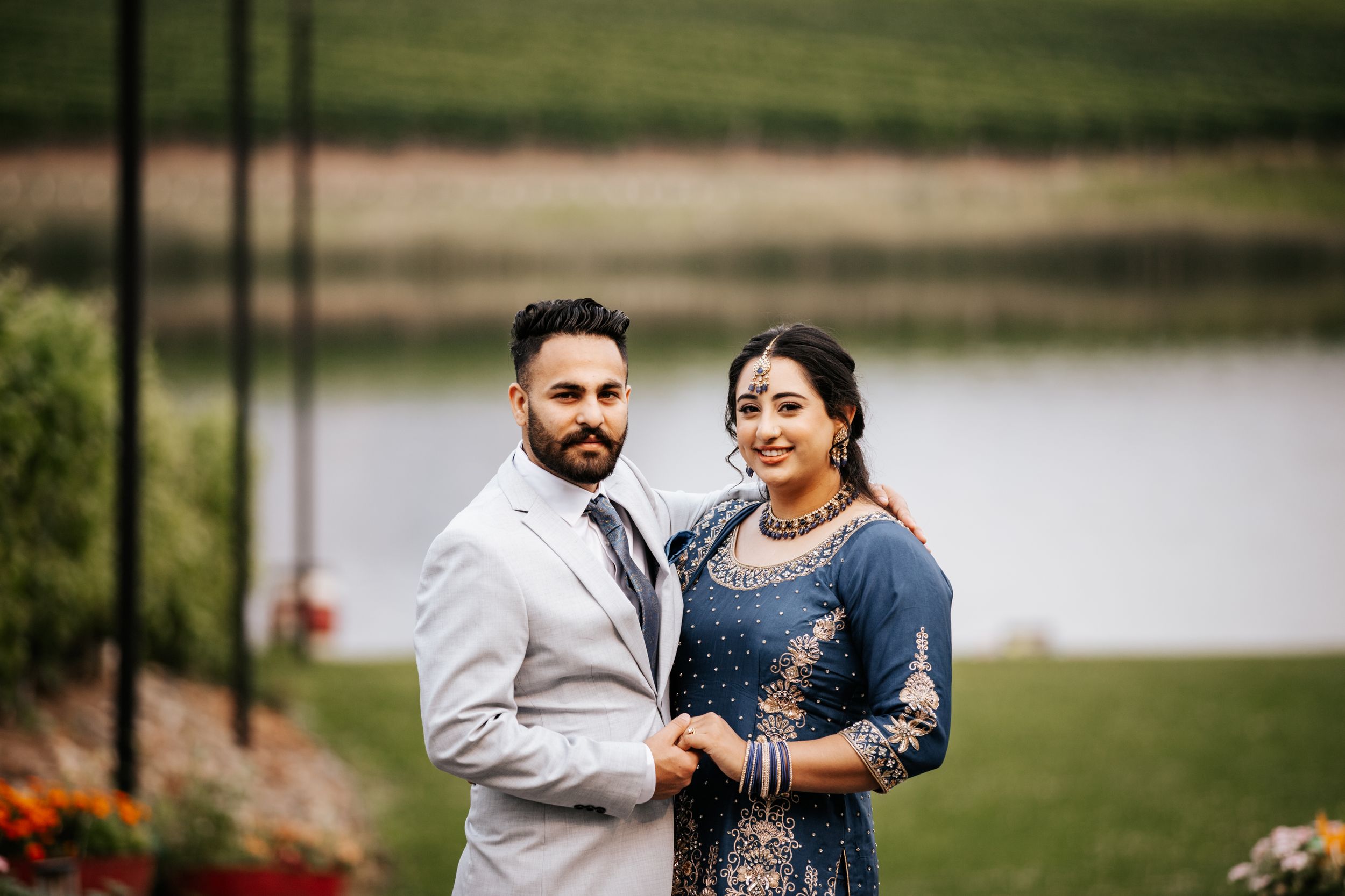 An elegant couple poses together outdoors near a lake, wearing formal Indian wedding attire.
