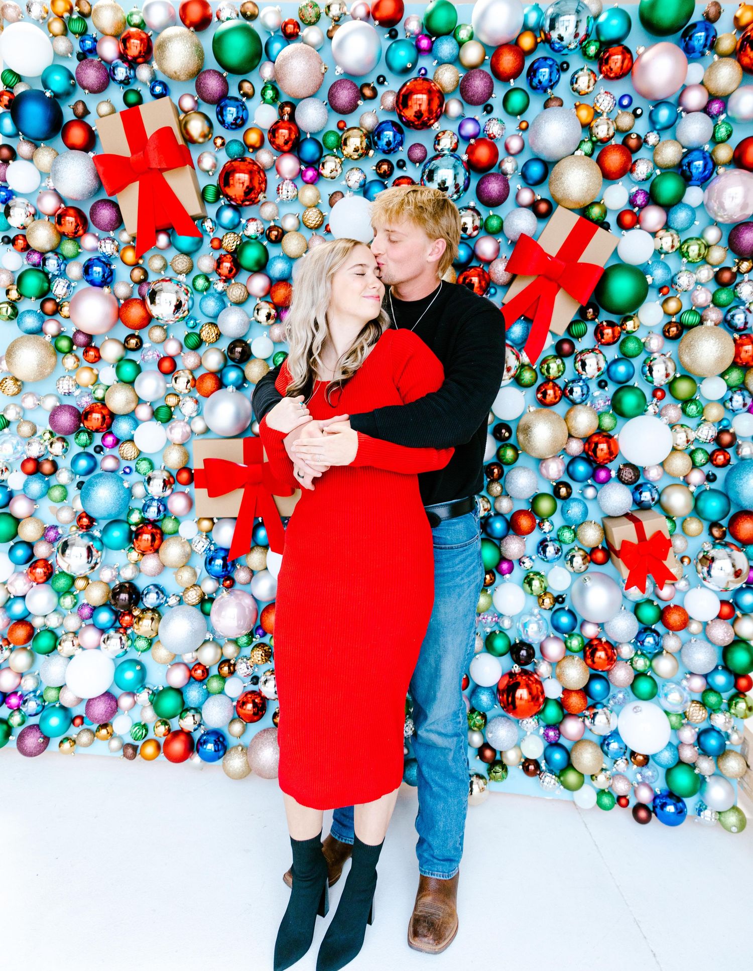 A couple embraces in front of a festive wall decorated with colorful Christmas ornaments and red gift boxes.