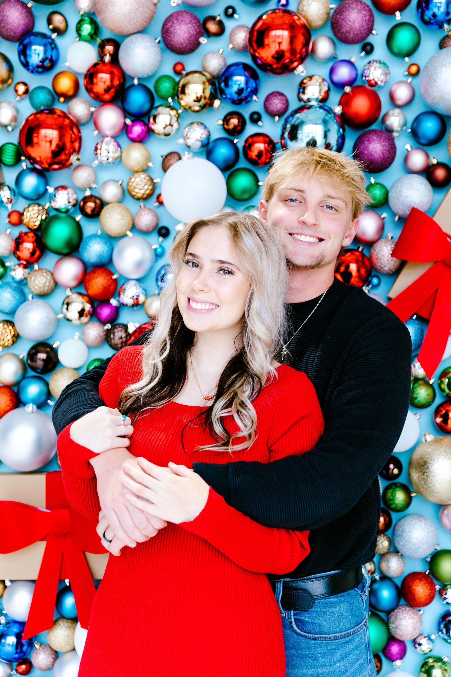 A couple poses in front of a colorful wall of Christmas ornaments wearing red and black holiday outfits.