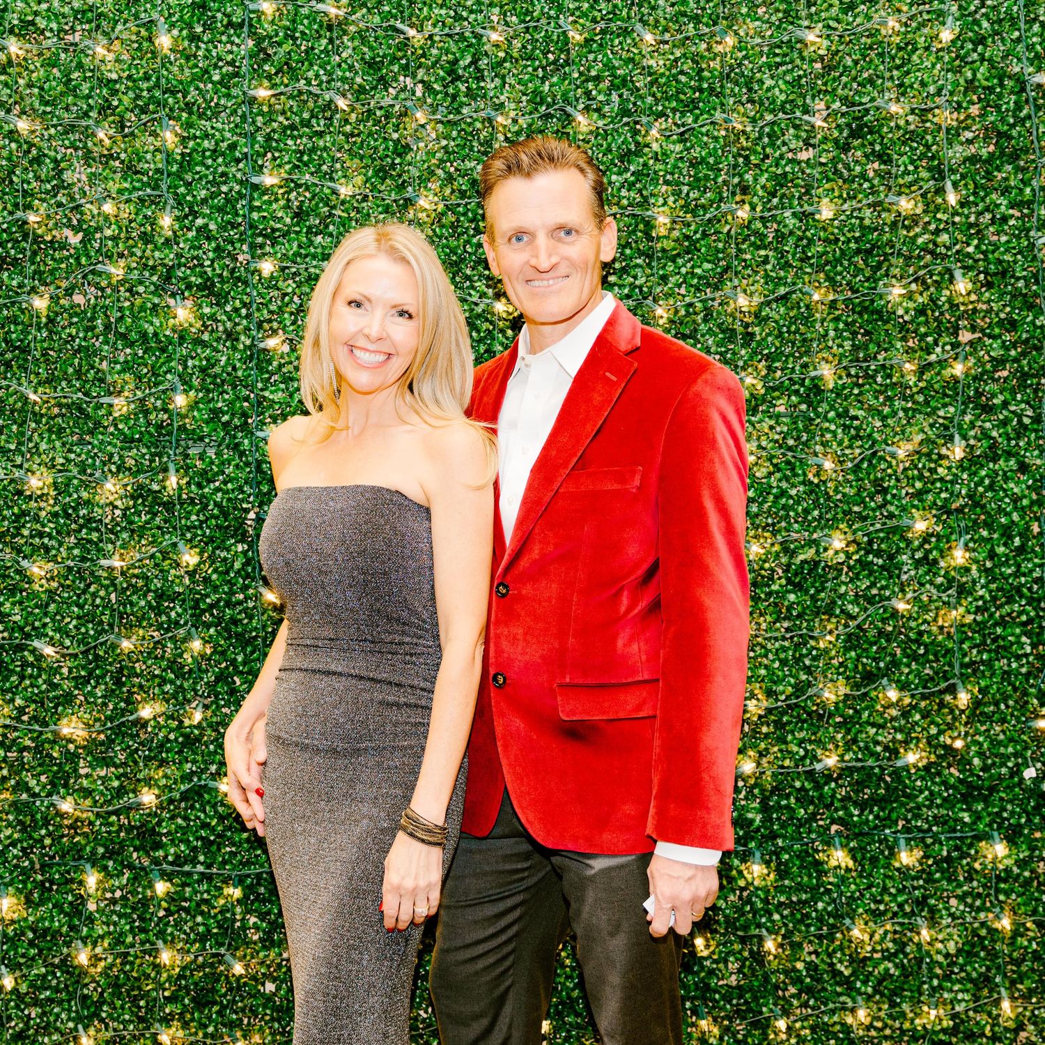Two people pose together against a green hedge wall with twinkling lights, wearing a gray dress and red velvet blazer.