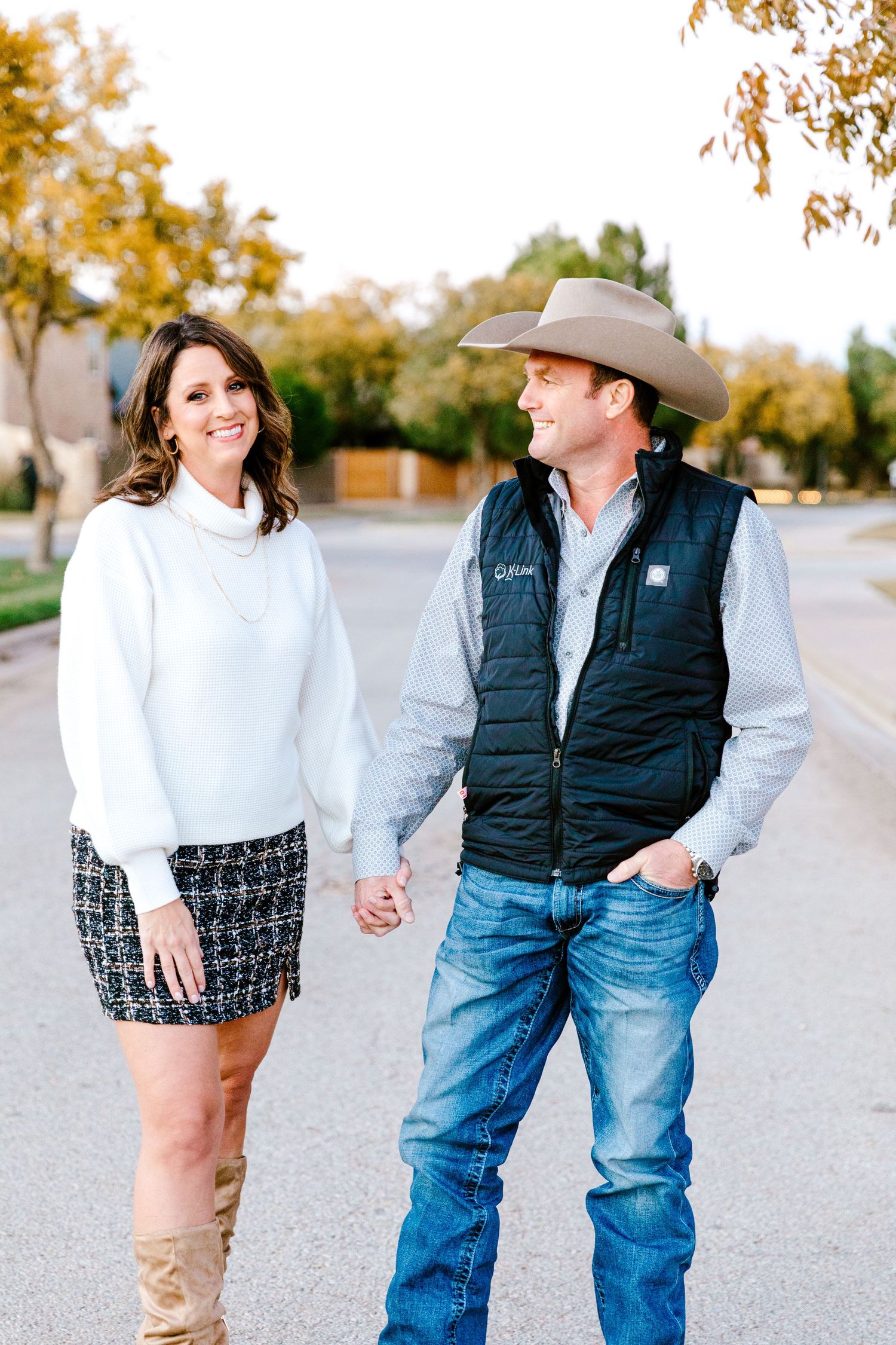A couple walking together on a street, with fall foliage in background, wearing casual western-inspired attire.