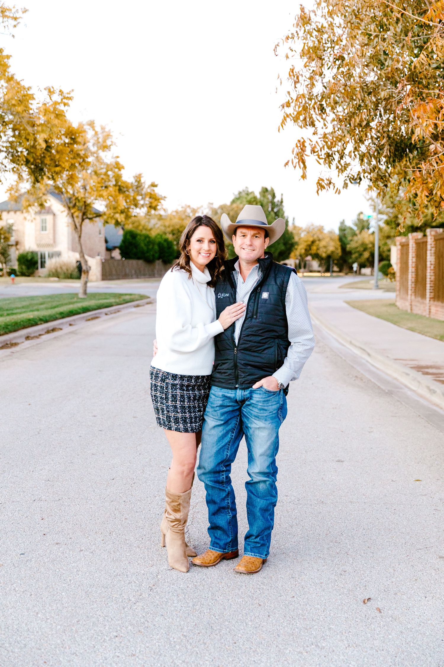 A couple stands together on a tree-lined street during autumn, wearing casual western-inspired outfits.