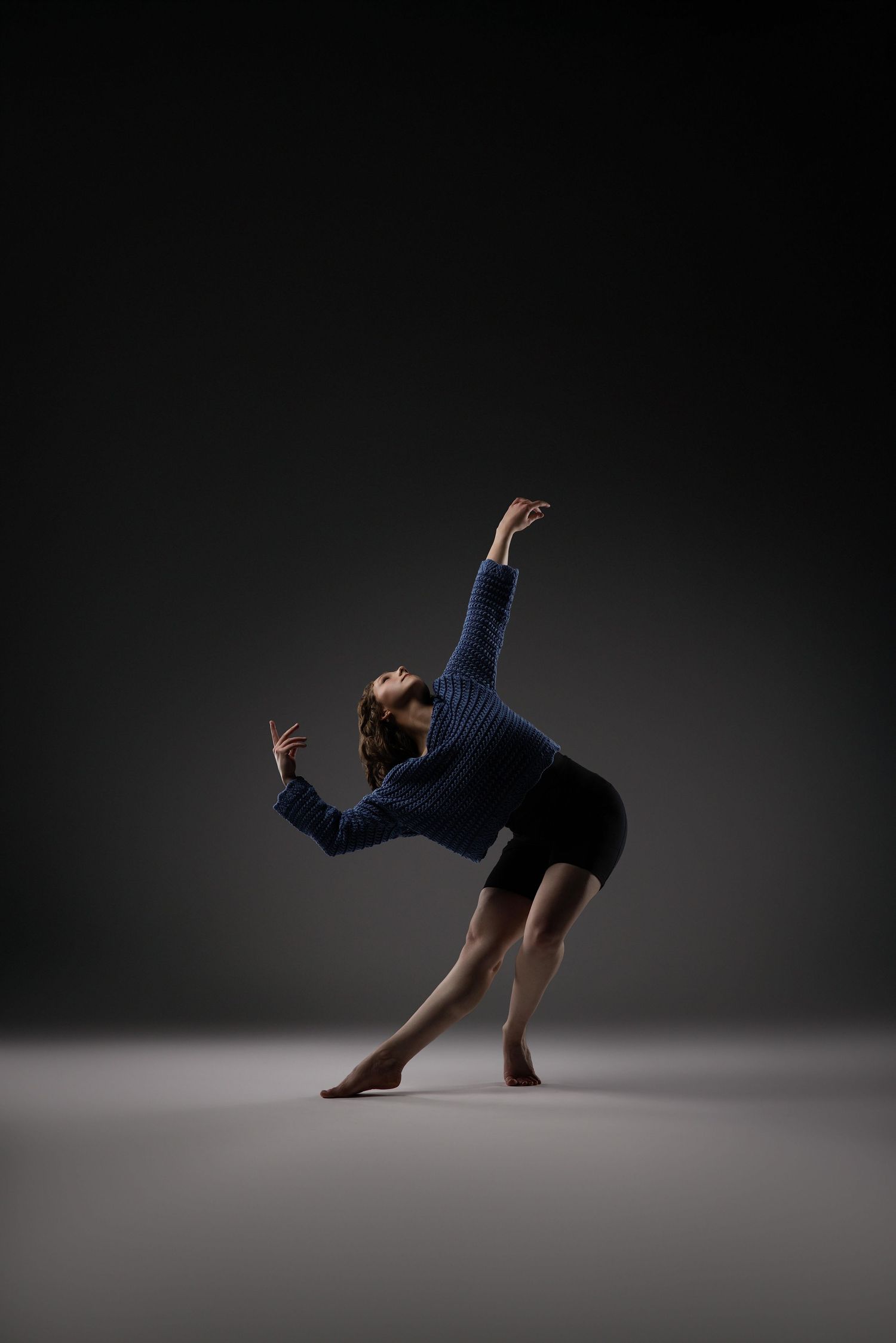 Graceful dancer in a navy sweater and black shorts performs expressive modern dance movement on dark stage.